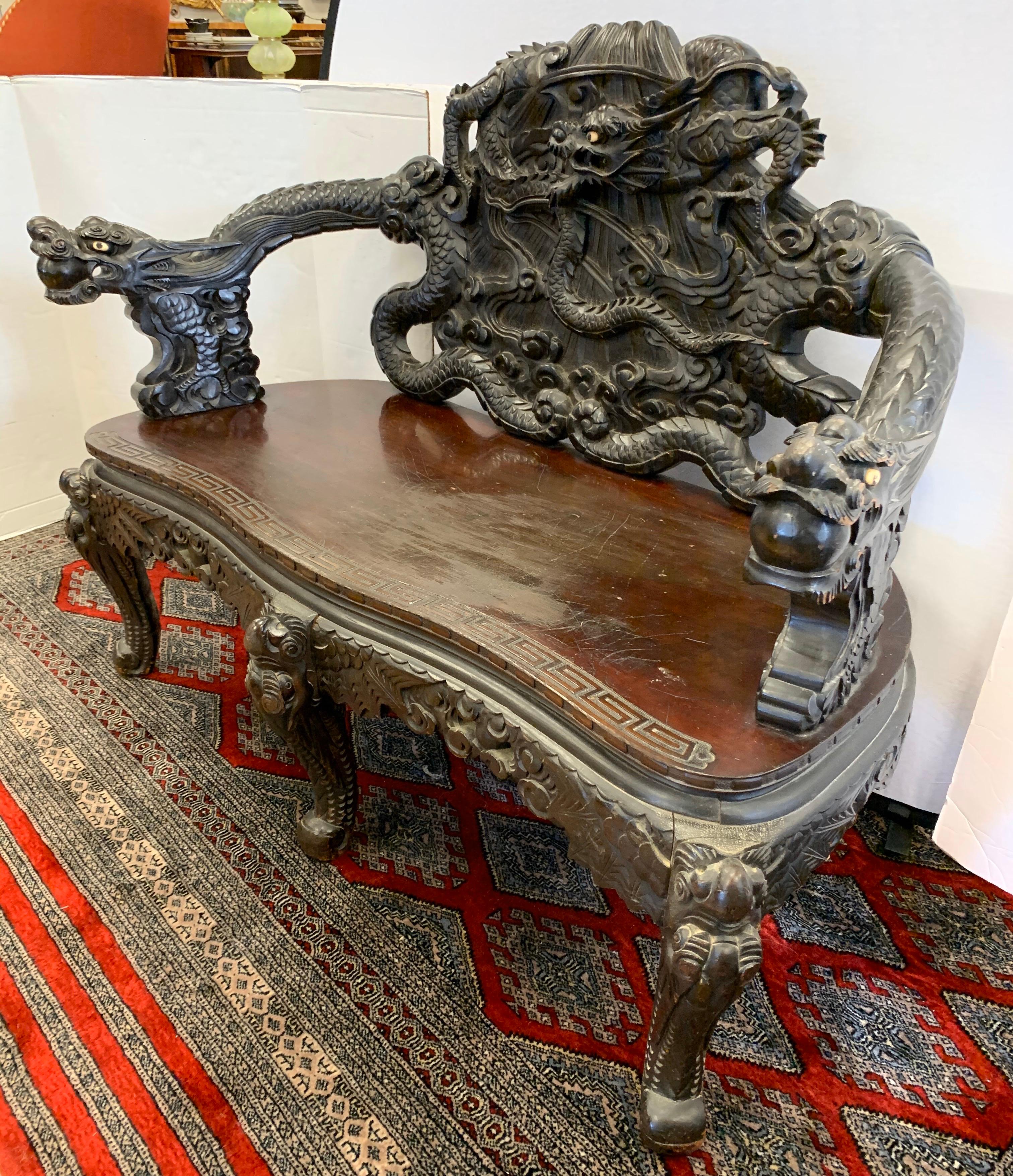 Antique and unique Chinese bench is finely and ornately carved with dragons with white eyes of hard stone. Center dragon is writhing and entwined within itself. Dragons on arms are holding pearls in their mouths, a metaphor for longevity and