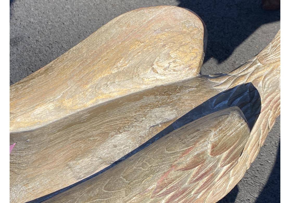 A large scale carved eagle with its wings closed as it reaches outward.  Its feet are perched on a scrolled detail on a block form base. With fine stylized carved feathers and eyes and traces of the original paint with green and red on the base and