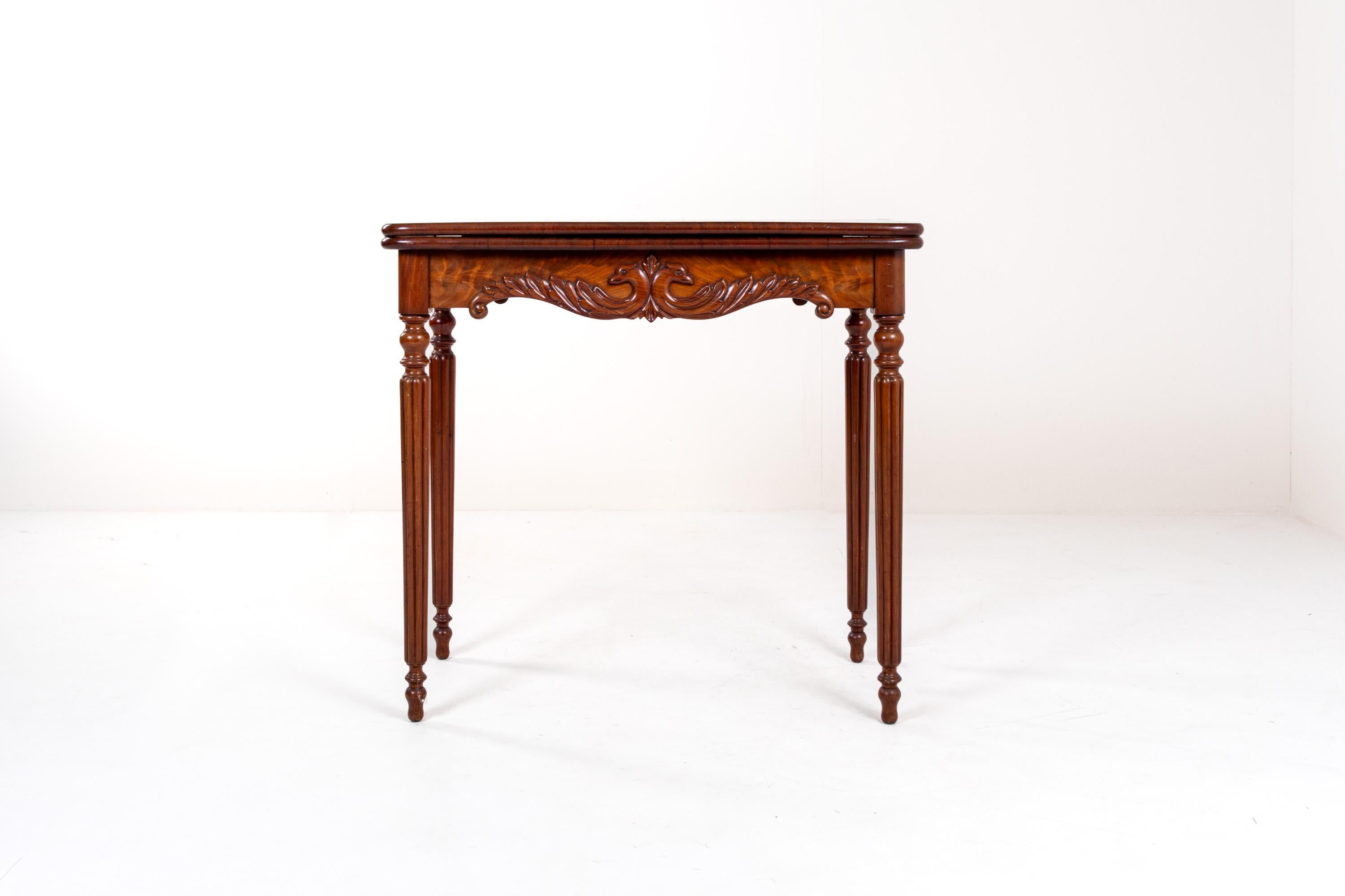 Table à cartes ou à jeux pliante, élégante et finement travaillée, probablement en placage d'acajou à la patine riche et chaleureuse. Le tablier présente des motifs feuillagés sculptés à la main et deux têtes d'oiseaux stylisées, typiques de