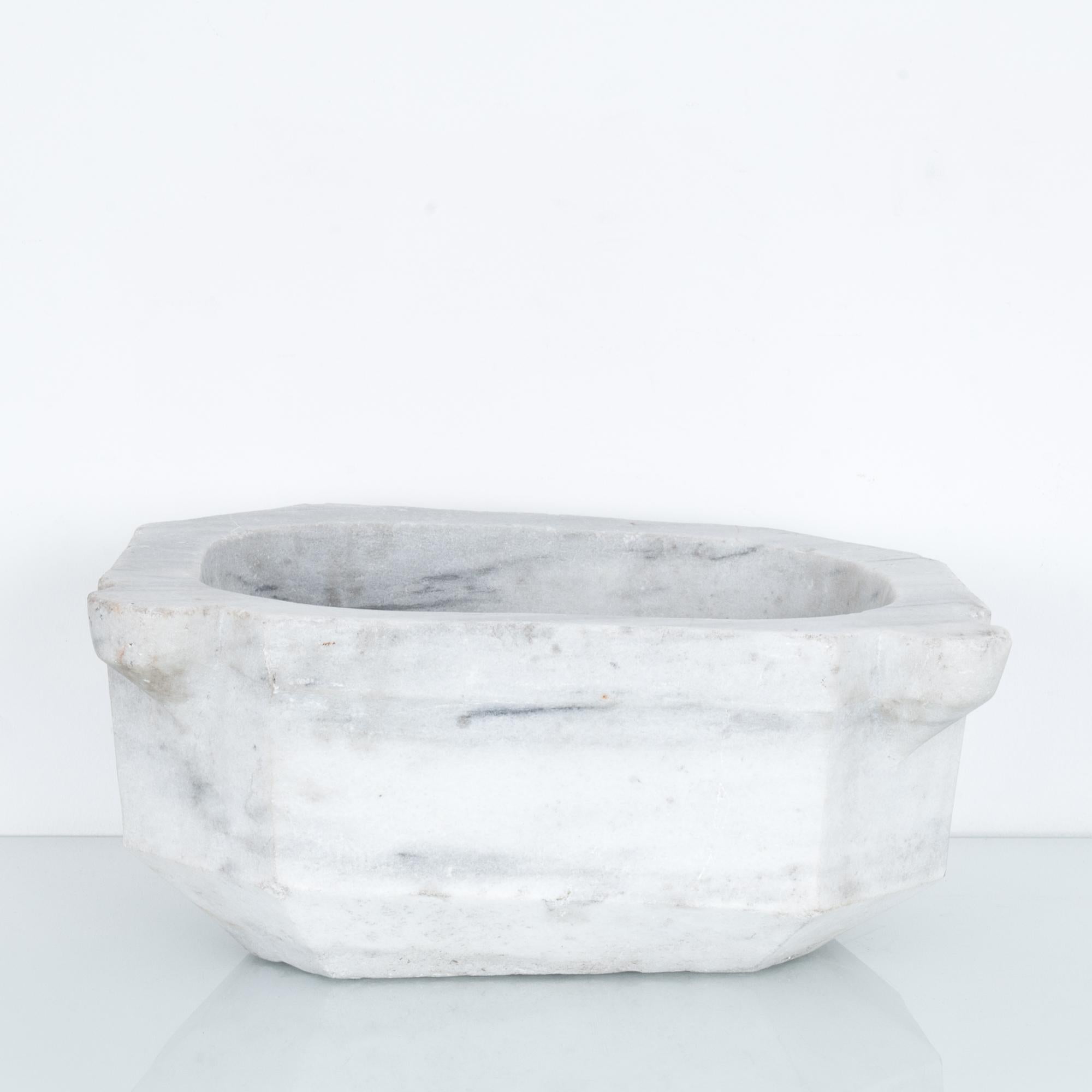 A stone architectural element, used formerly as a font or stoup. Found in ecclesiastical architecture, this basin for water would be placed at the entrance to a church. Rendered in cloudy grey marble, with a rustic textured finish, this stone object