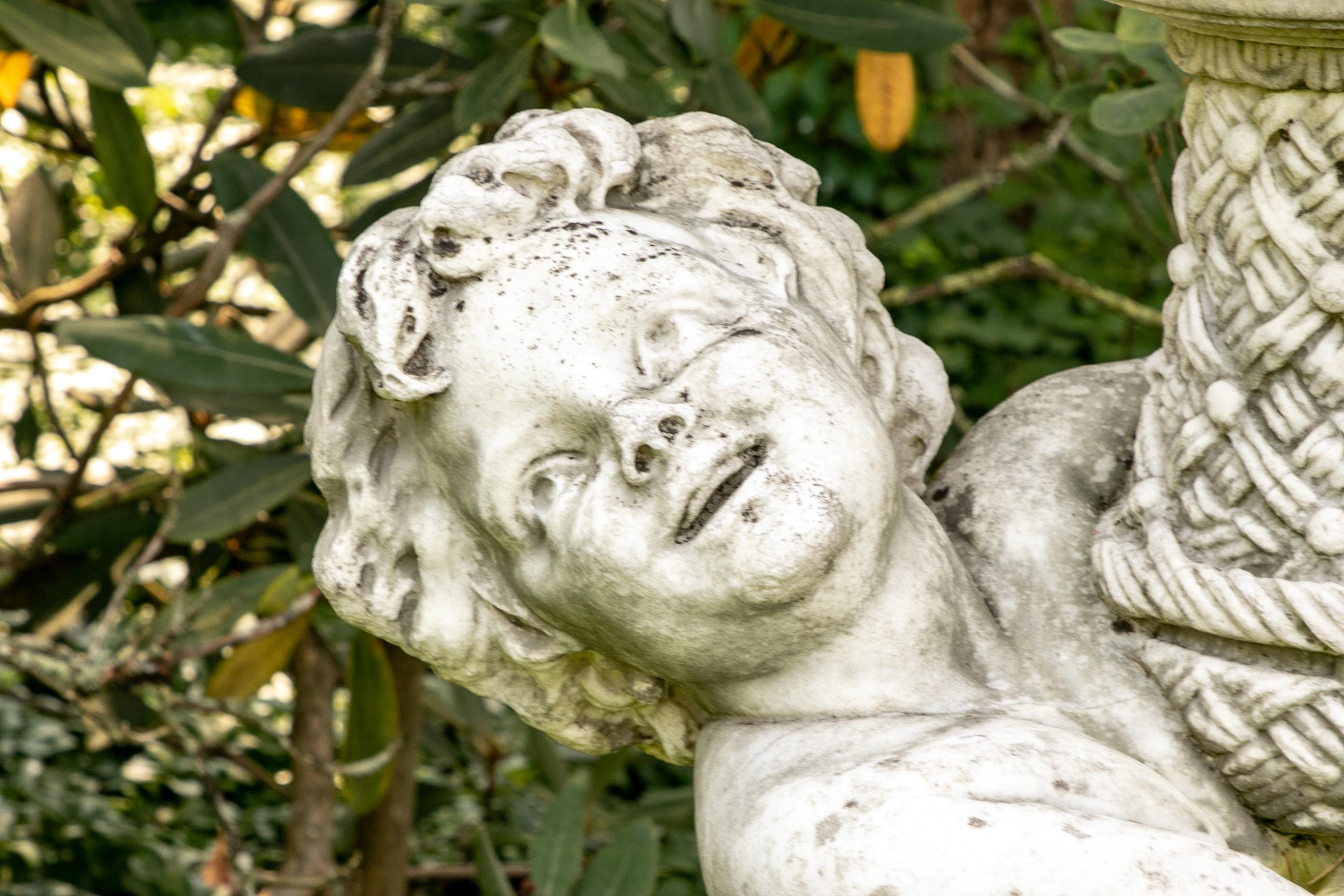 Eine großformatige Skulptur eines lachenden Putto, der auf einem kunstvoll beblätterten Sockel steht und eine Vase mit Korbgeflechtfuß trägt, die mit großen Akanthusblättern (einige mit kleinen Bohrlöchern) überquillt. Der quadratische Sockel mit
