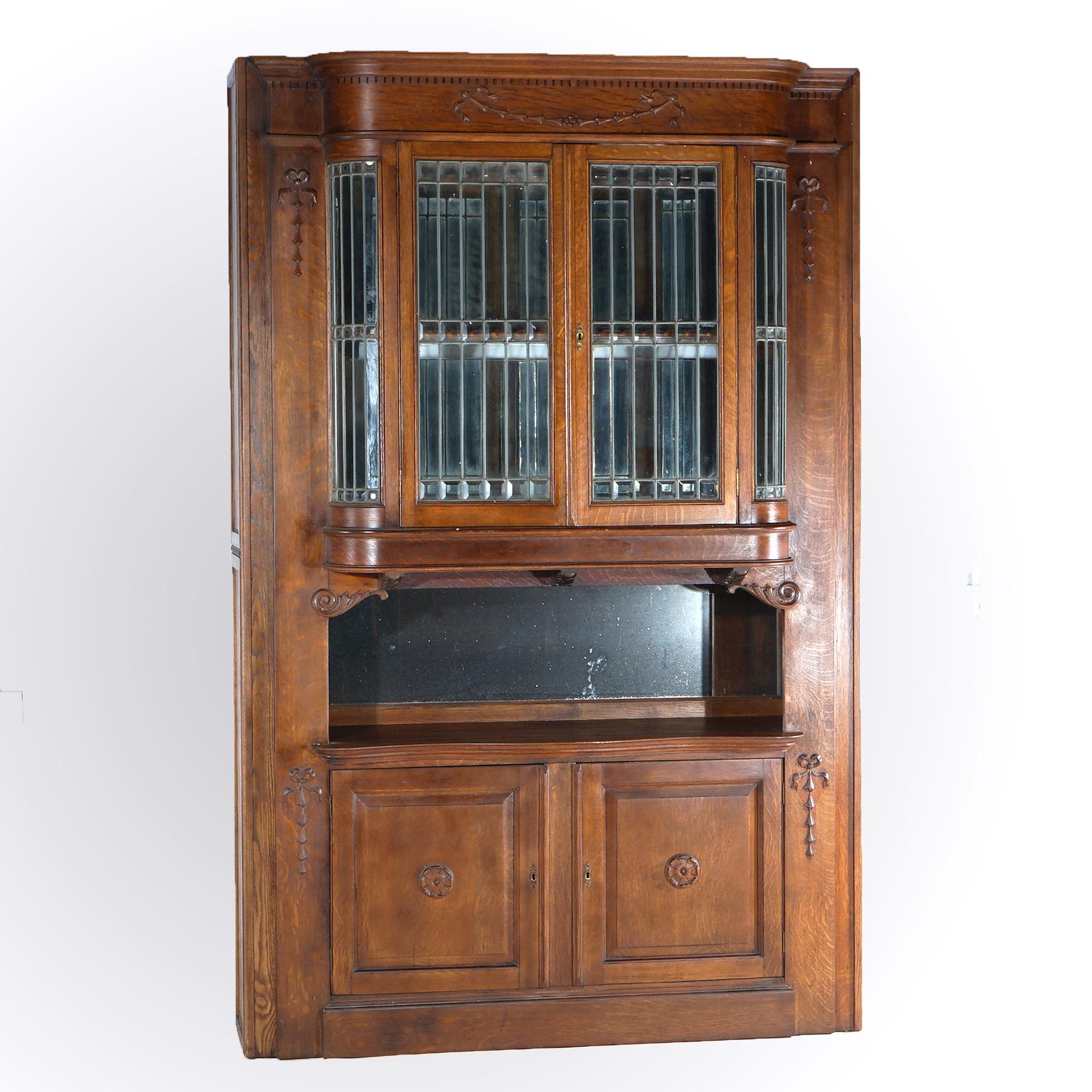 An antique buffet sideboard offers oak construction with upper china cabinet having double curved leaded glass doors opening to shelved and mirrored interior over double blind cabinet base; carved inverted bellflowers, rosettes and scroll elements;