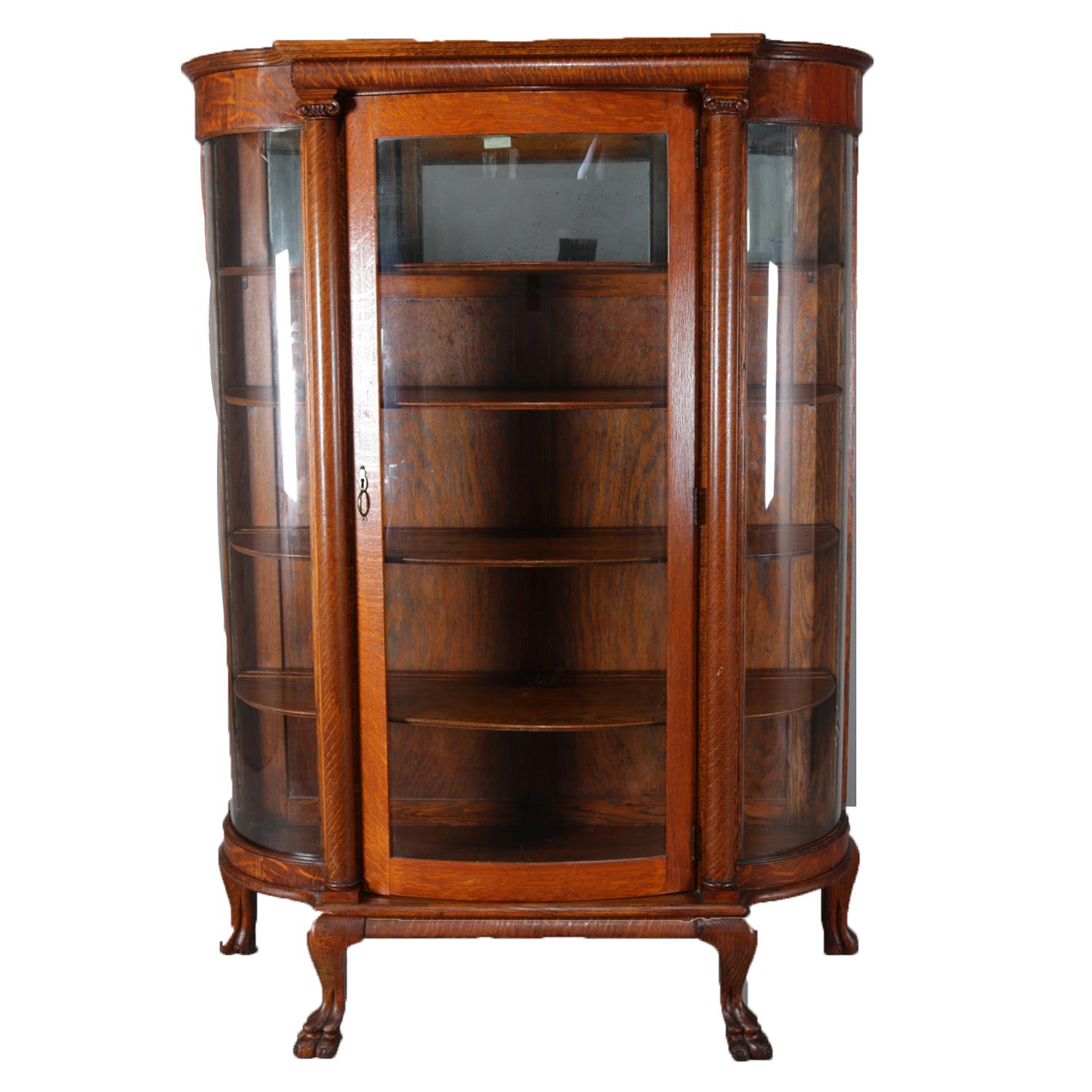 An antique Victorian china cabinet features oak frame with single glass door flanked by Corinthian column supports and curved glass sides, fixed shelf interior with mirrored upper, and raised on carved paw feet, circa 1910

Measures: 61.75