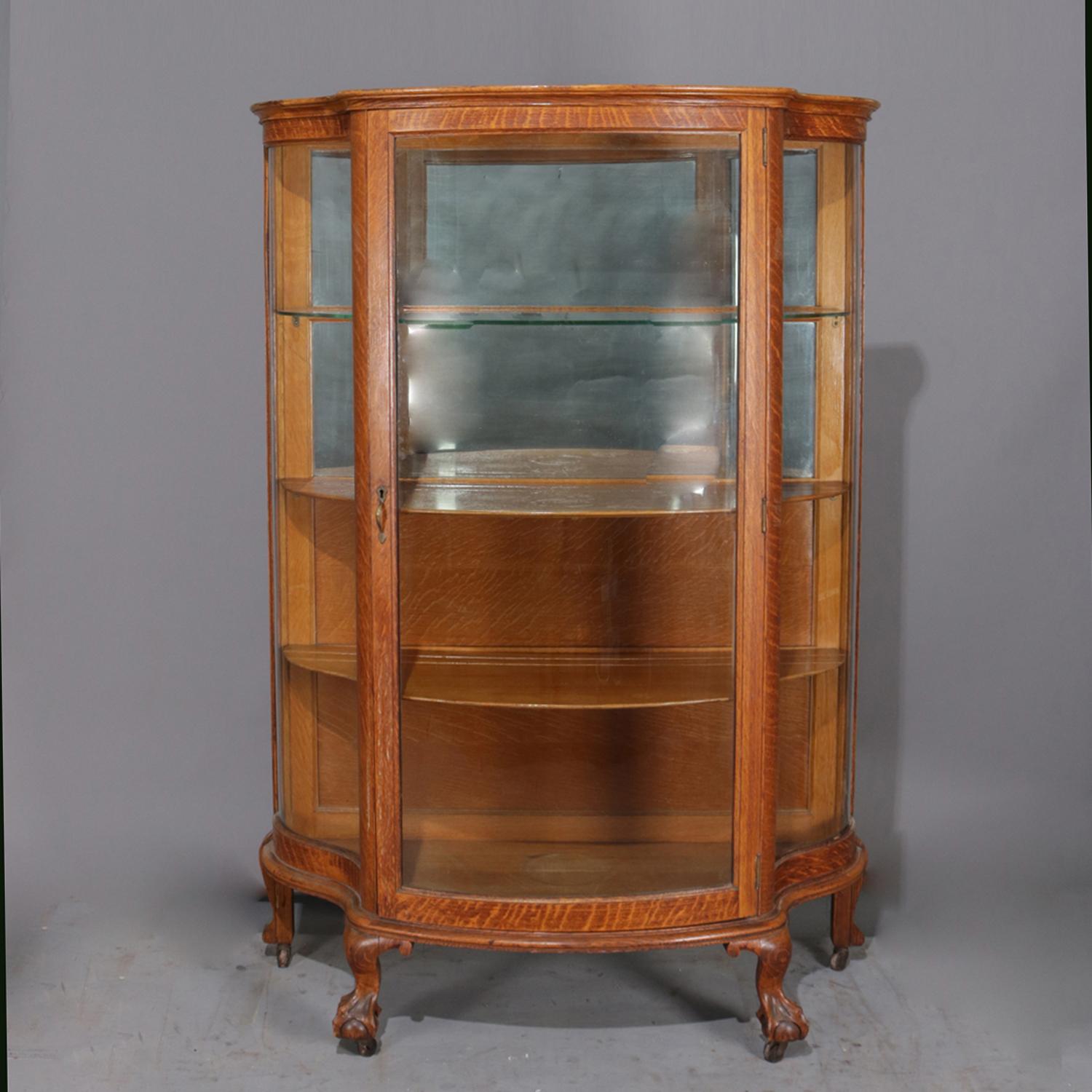 An antique china cabinet by R. J. Horner features quarter sawn oak construction in serpentine form with curved glass sides and single front access door opening to interior having upper with mirrored back and glass display shelf, lower with wood