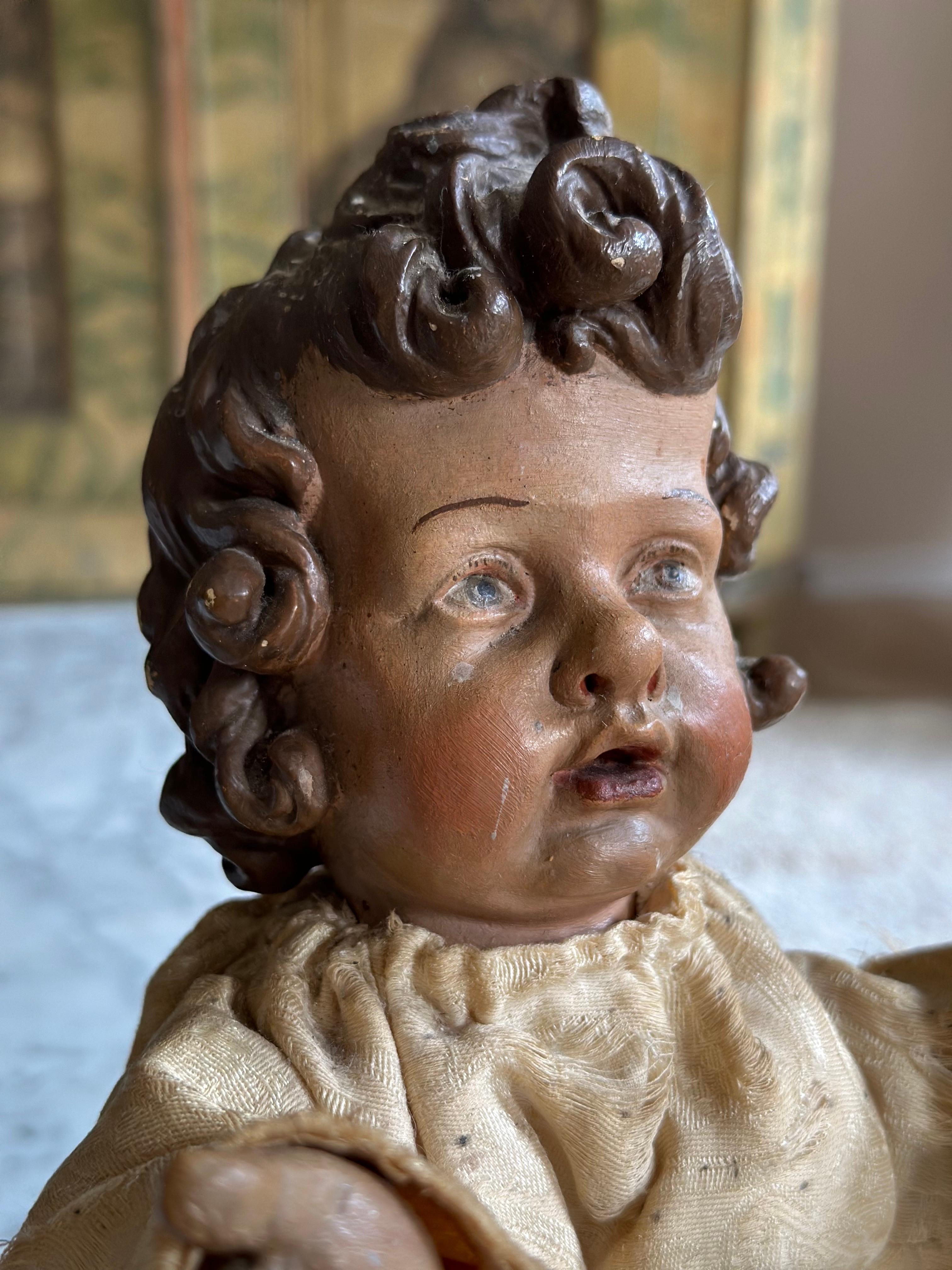 Cette figurine en bois sculptée et finement travaillée, datant d'environ 1800, présente une qualité artistique et un niveau de détail exceptionnels. La figure assise, vêtue de son vêtement blanc d'origine, dégage une présence douce et enfantine,