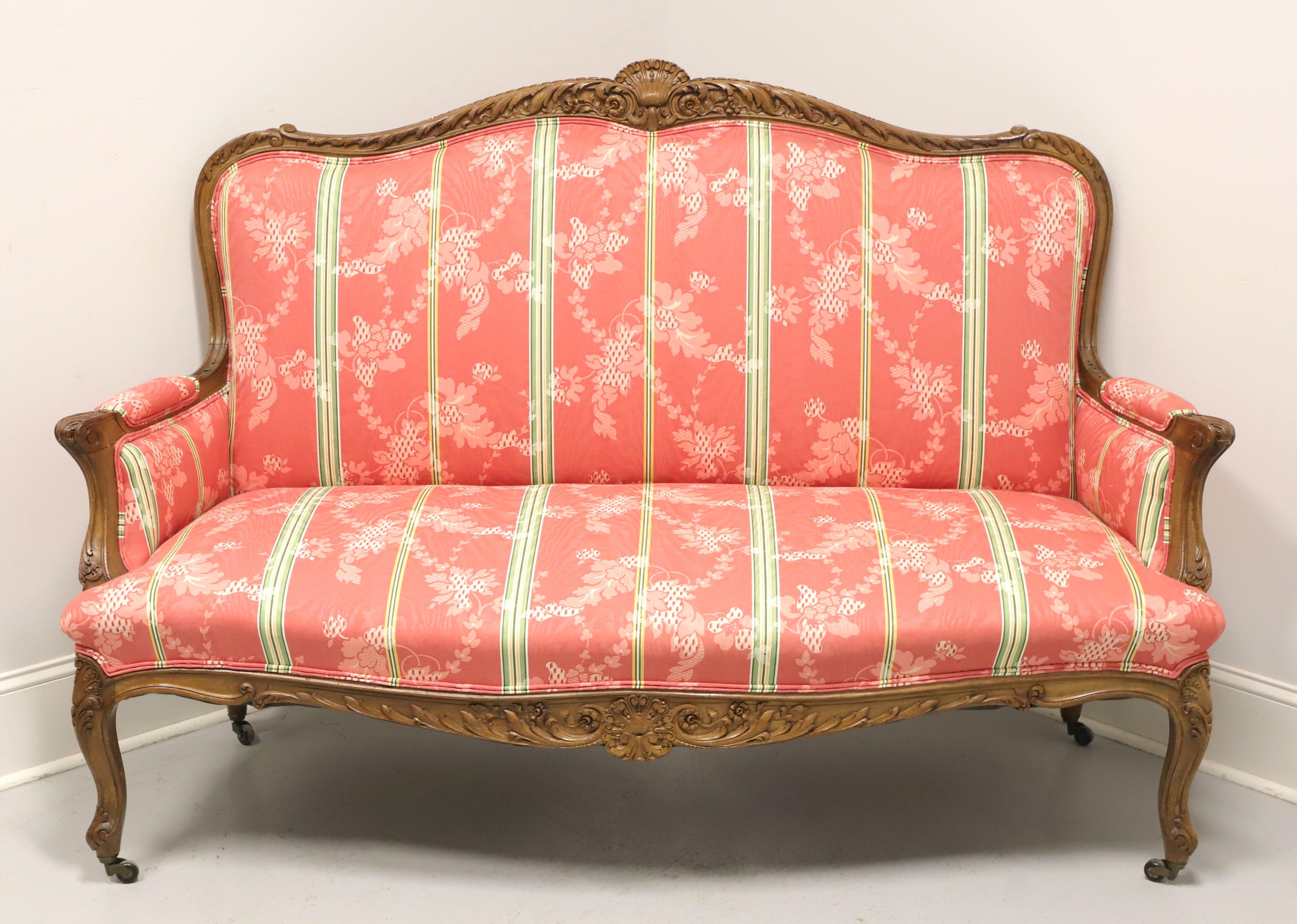 An antique French Country Louis XV style settee, unbranded. Frame made in the 19th Century of finely carved walnut with high back; decorative carving to back, arms, apron & knees; fabric upholstered arms; and curved front & back legs with metal &