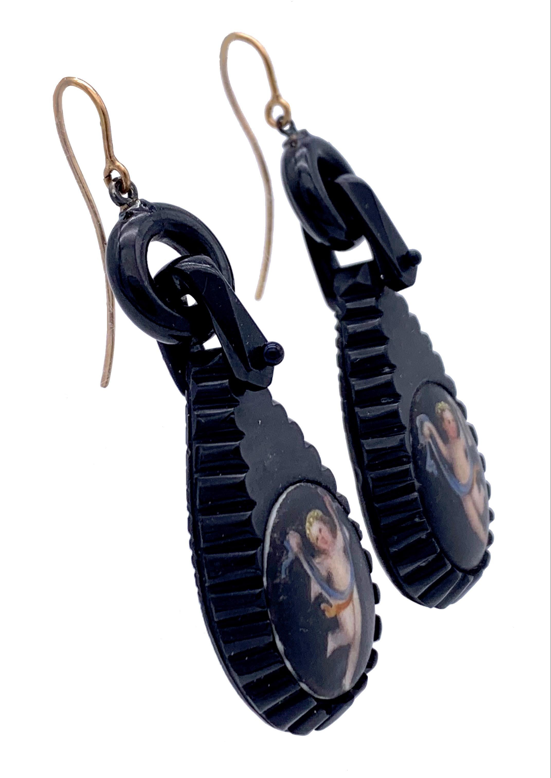 These substantial carved Whitby jet earrings are decorated with painted porcelain plaques depicting dancing cupids holding garlands between their hands. The little cupids are wearing wreaths on their heads.