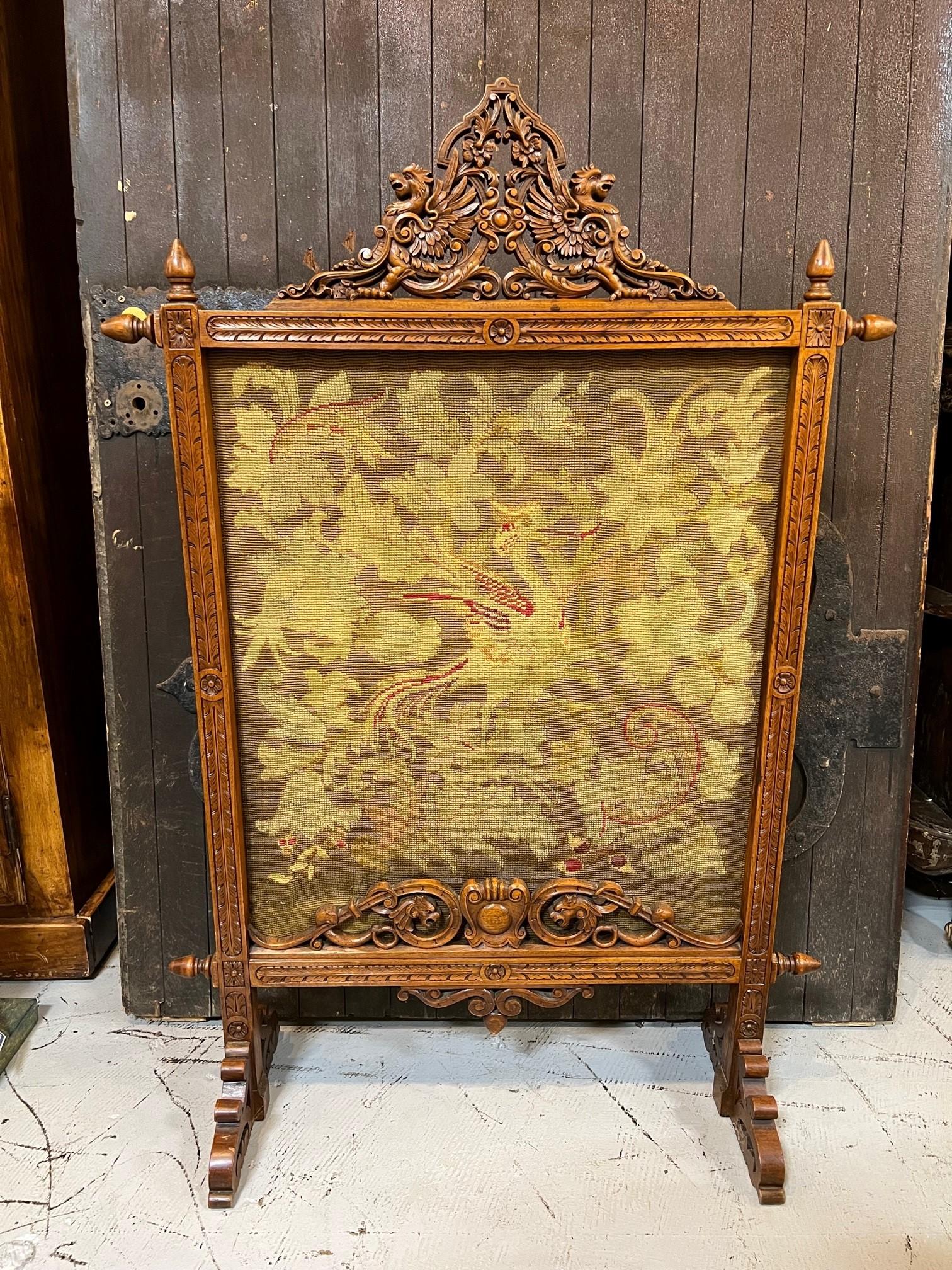 Beautiful late 19th century carved wood fire screen with a center crest of winged griffons and the original needlepoint tapestry center. The carved wood frame is made of walnut and is supported by scroll carved wooden feet which are very sturdy. The