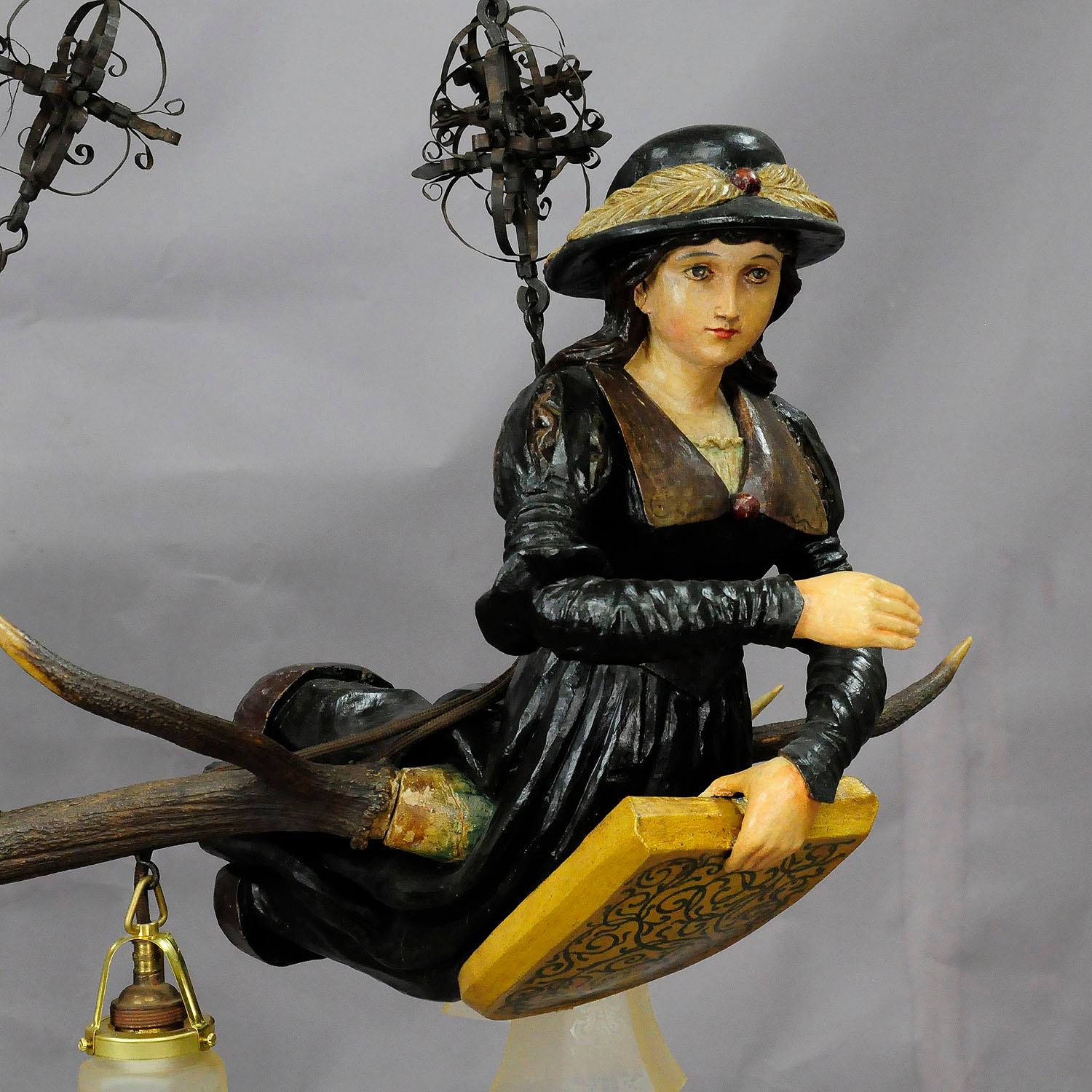 A wooden hand carved and painted statue of a Victorian lady fixed on a large pair of original stag antlers. named Lusterweibchen or Lüsterweibchen, executed circa 1920 with hand forged iron suspension and 4 electrified fittings with glass shade,