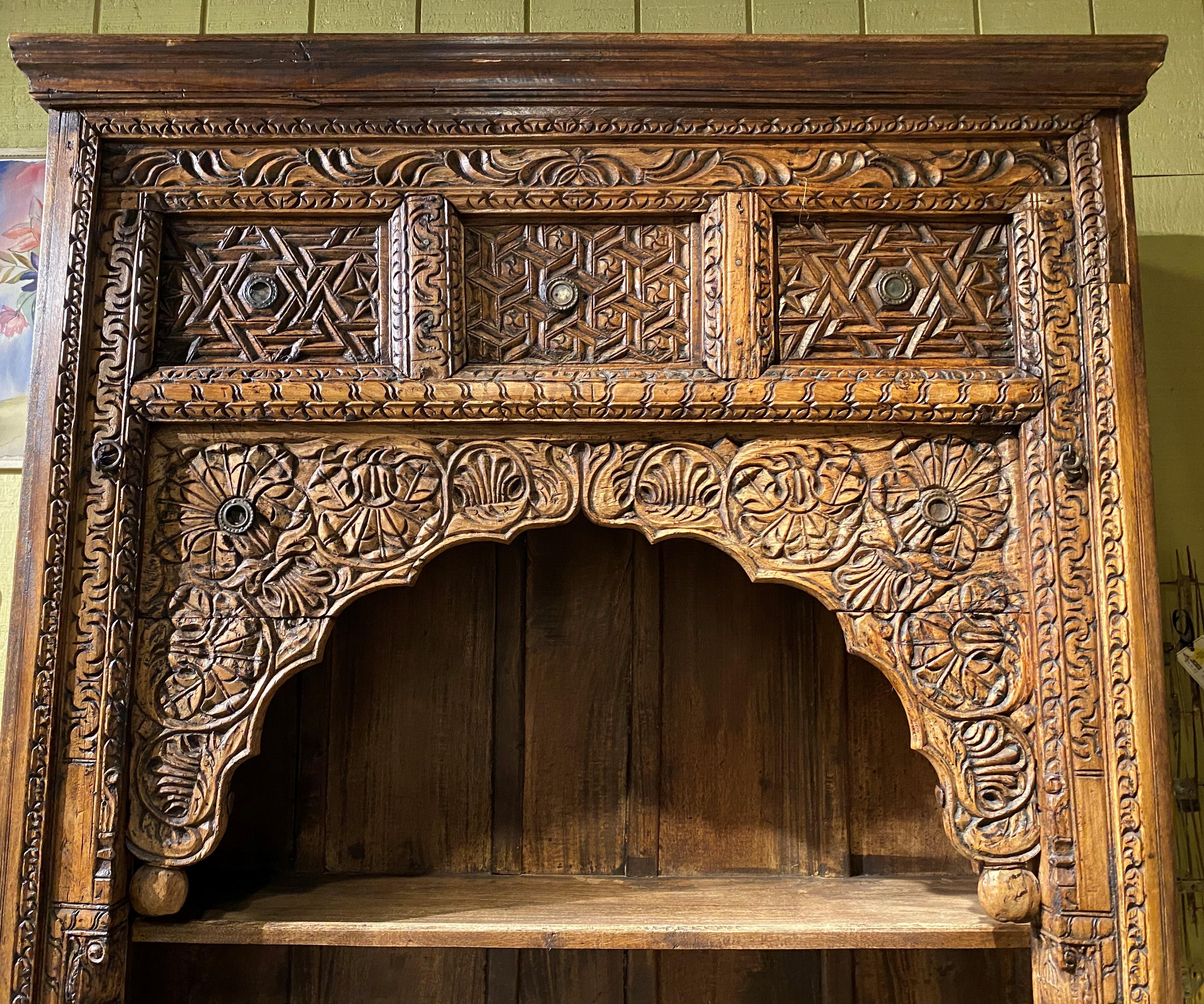 Antique carved wooden Indian arched doorway dating to the late 19th or early 20th century that has been converted into a three- shelf bookcase, with a molded cornice and upper carved panels adorned with metal decoration. Very good overall condition,