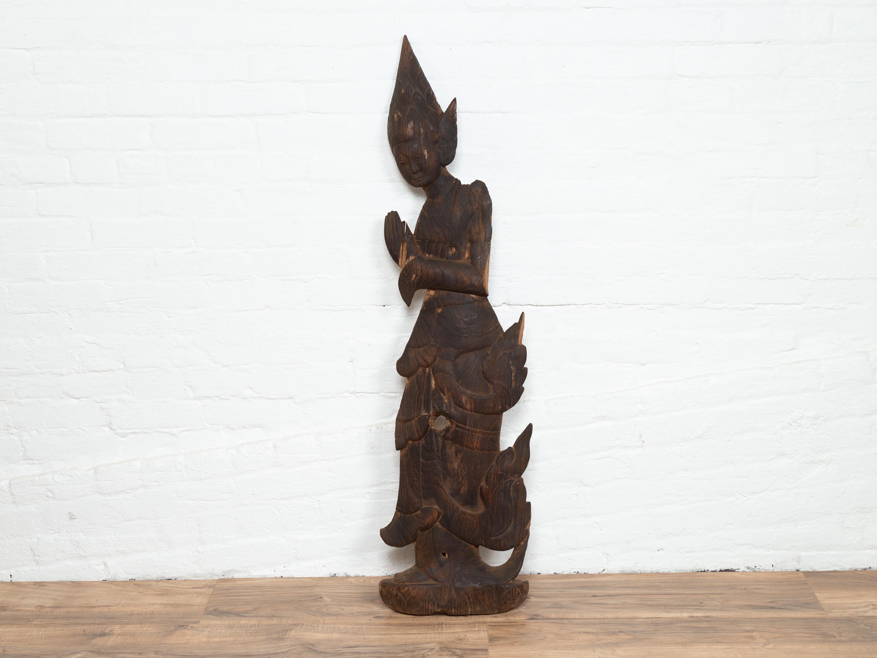 A carved Thai wooden sculpture of a praying Apsara, with flat back and dark patina. Attracting our attention with its dark patina and angular depiction, this wooden sculpture, unfinished and flat on the back depicts a praying Apsara, a female spirit