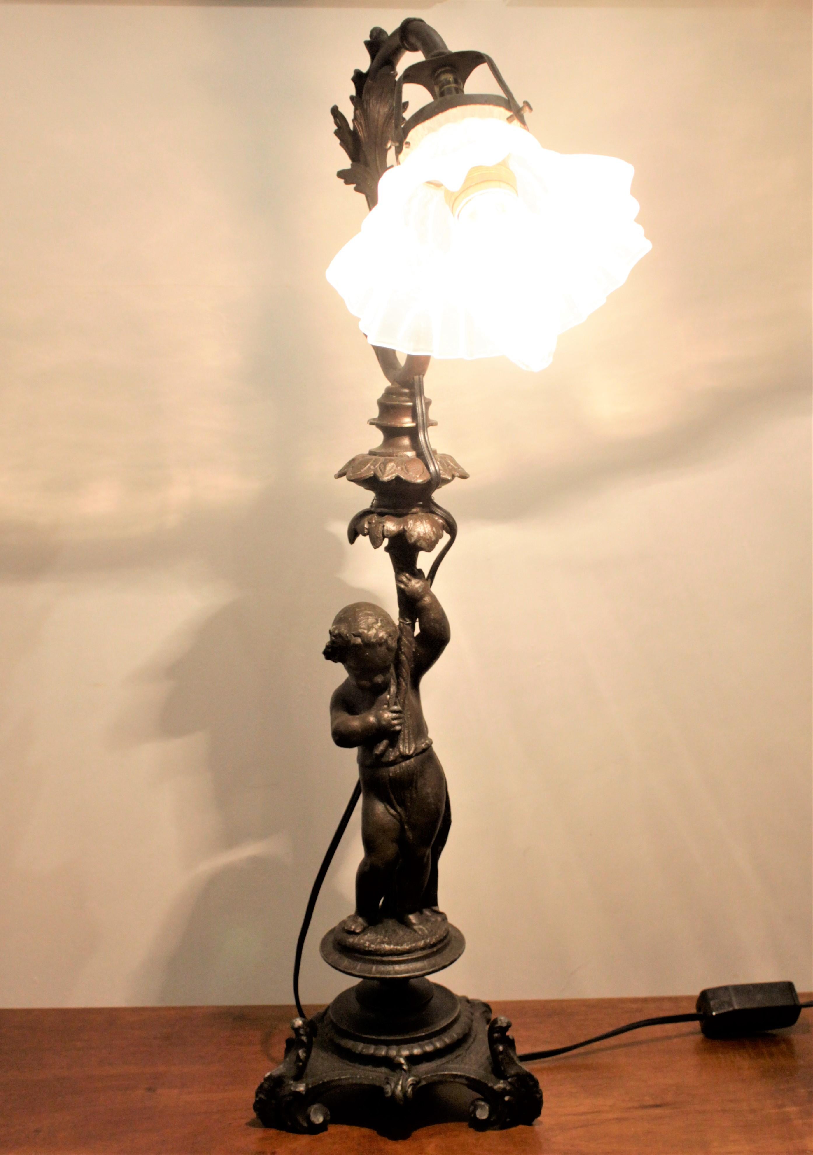 This antique cast spelter figural cherub lamp has no maker's marks, but is presumed to have originated from Italy in the late 19th century and in Victorian style. The lamp features an ornately cast cherub standing on a round pedestal which sits on a