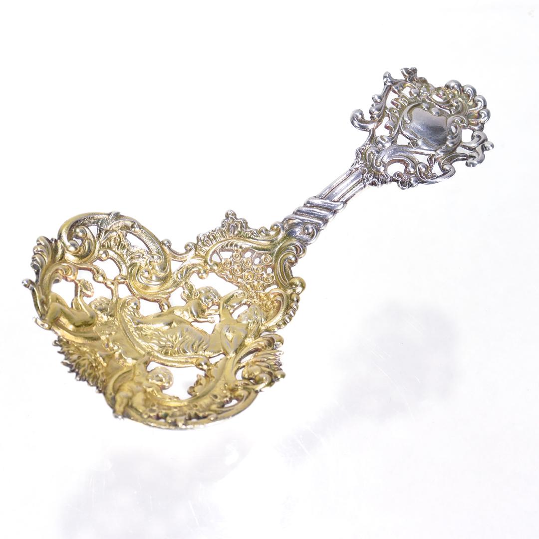 A fine, antique bon bon spoon.

By Gorham.

In cast sterling silver. 

No. 589.

With an ornate, reticulated handle and a gilt, pierced bowl with a scene including a bare-breasted woman and 3 cherubs or putti. 

Simply a wonderful Gorham