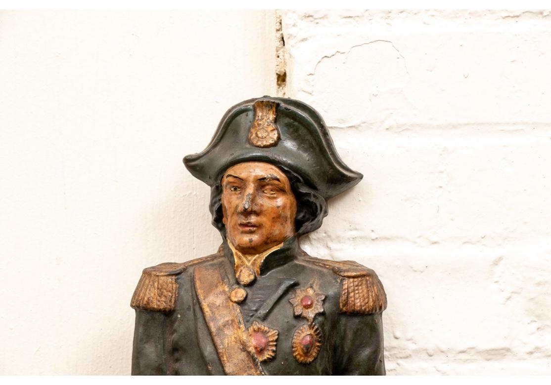 Antique painted iron plaque depicting a standing Admiral Nelson clutching a telescope in his only hand while standing next to an anchor wrapped in rope to one side and an anchor to the other. Below his feet is a decorative plinth comprised of two