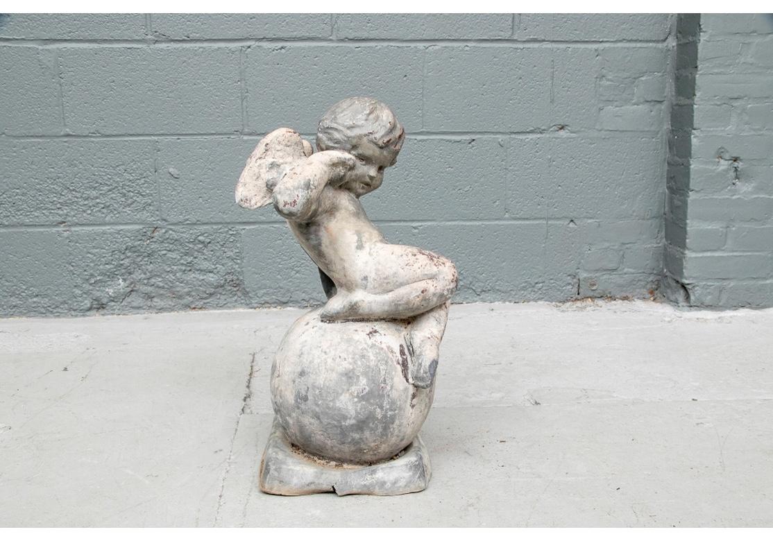 The Winged Angel sits curled up on a sphere. He raises one hand to his ear as if to listen to a seashell. Originally painted in brown and off white. 
Measures: Height 20 1/2', Width 11