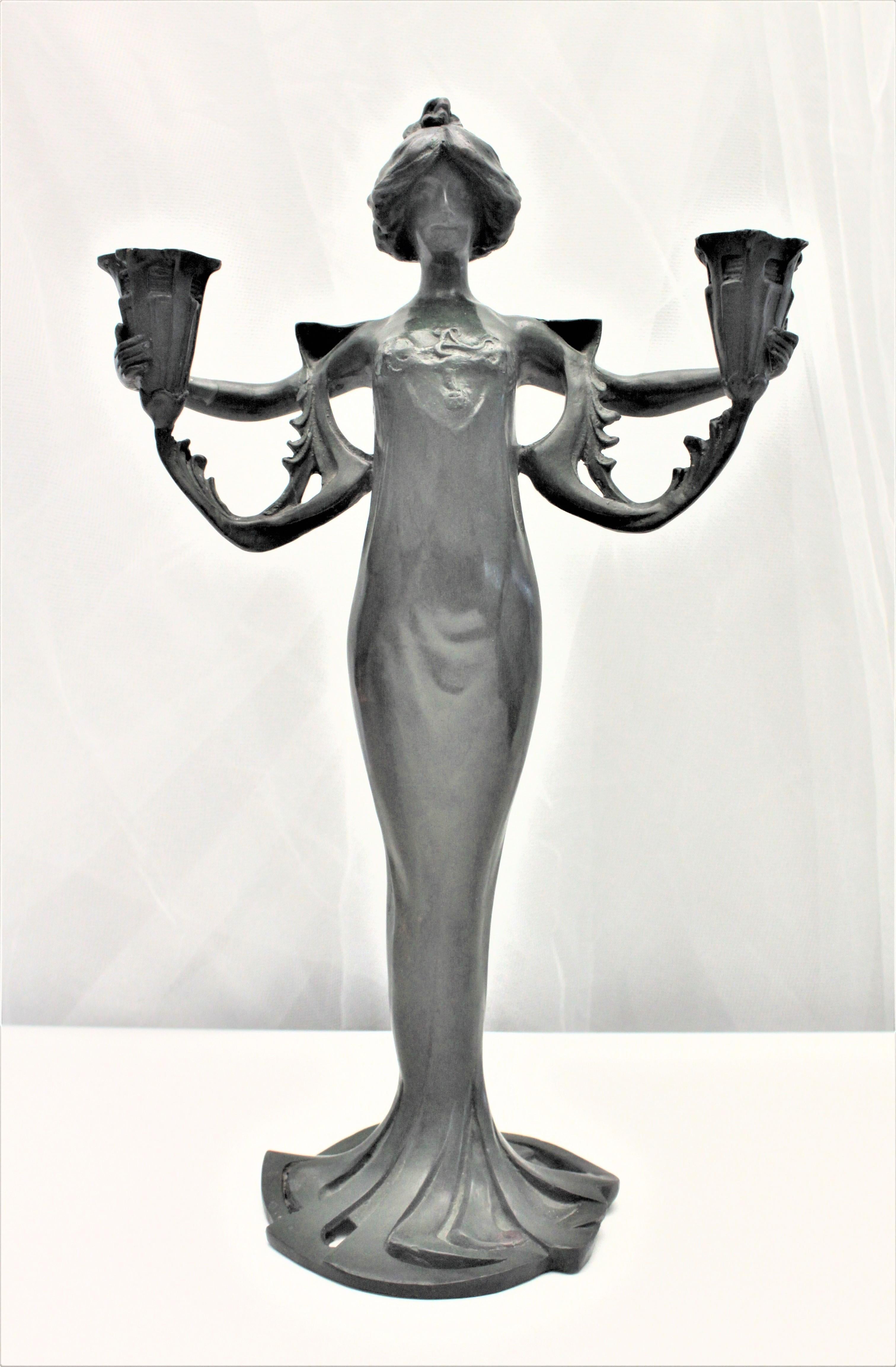 This antique candleholder is unsigned and unmarked, but believed to have been made in the United States. The cast spelter candleholder depicts a robed woman with outstretched arms which function as a double candleholder. This stylized female cast