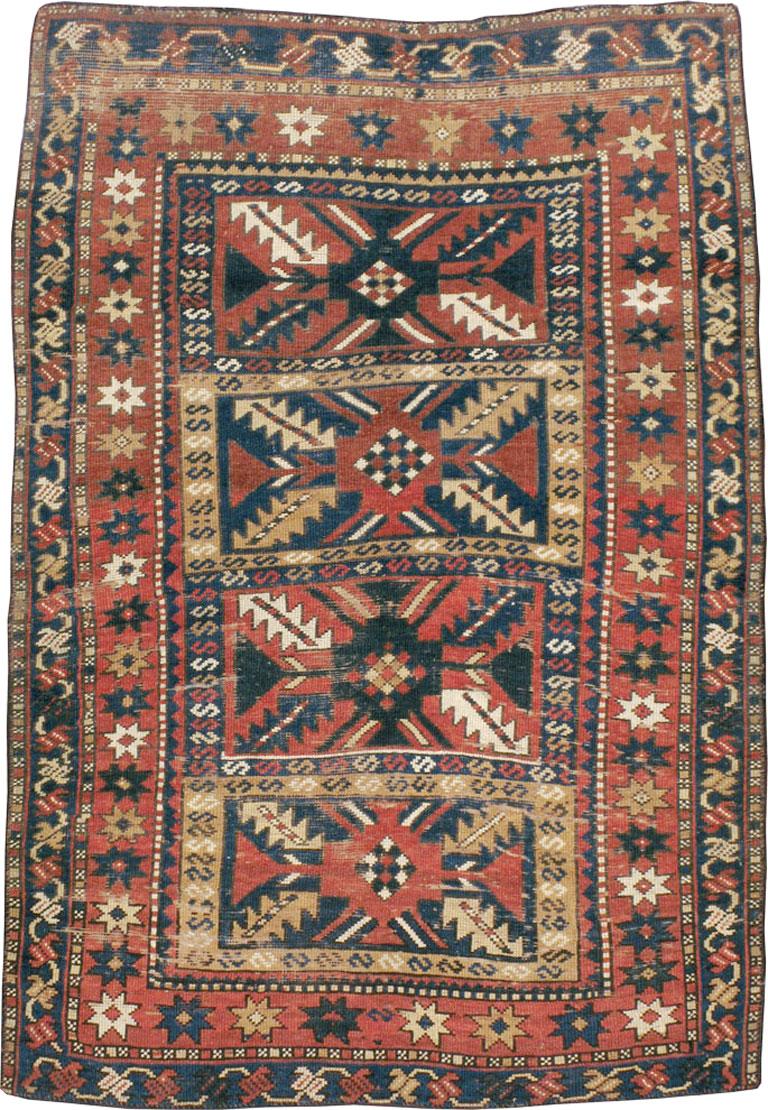 An antique Caucasian Kazak rug from the early 20th century. The warm madder red field is divided into four rectangular panels, each with a central motif radiating four serrated leaves. The rust main border displays disjoint stars while the dark blue