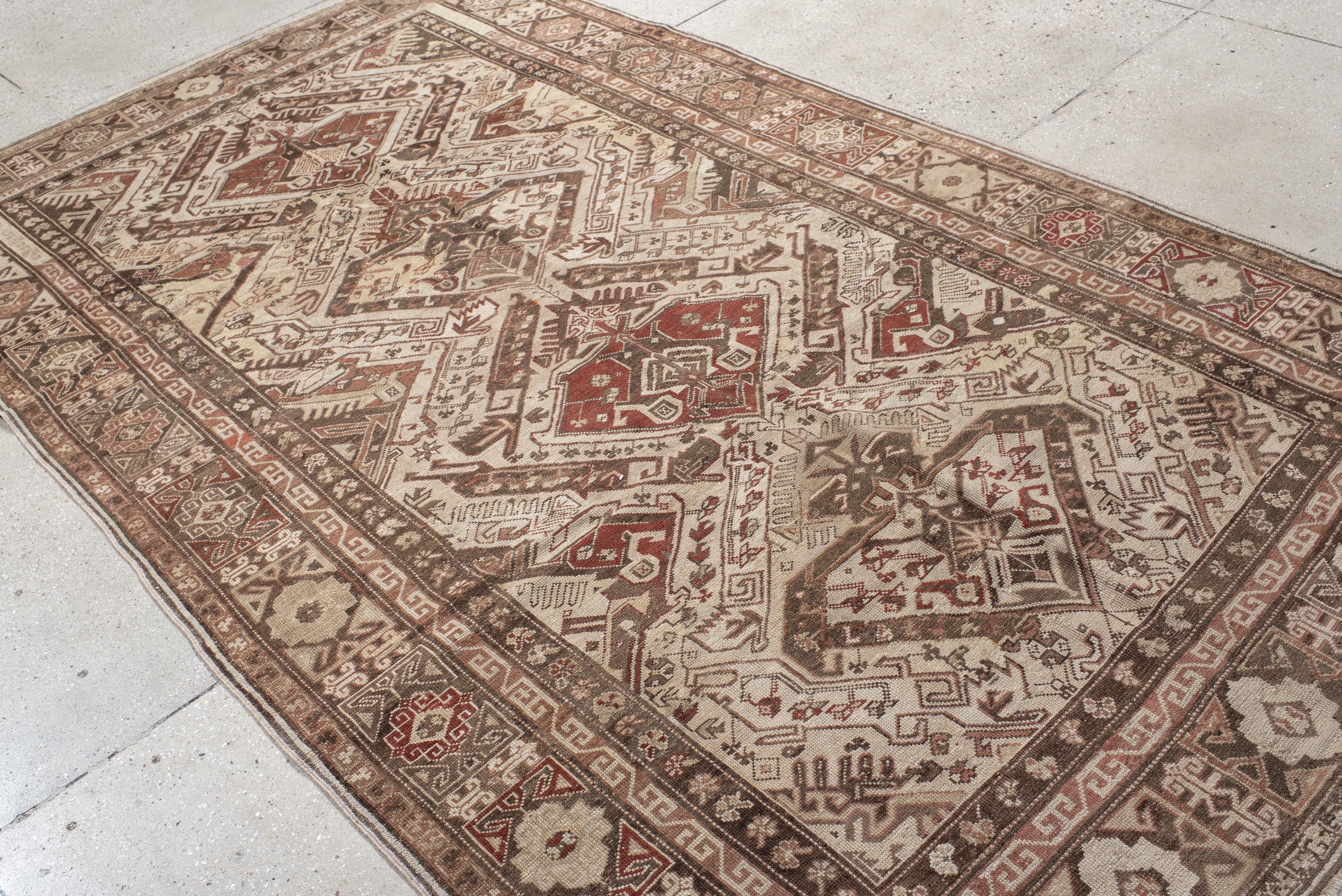 Ce tapis caucasien ancien présente une série de médaillons en escalier, aux couleurs rouge brique, camel et moka, sur un fond ivoire. La composition est audacieuse et graphique, avec des motifs tribaux angulaires, des éléments animaux et des