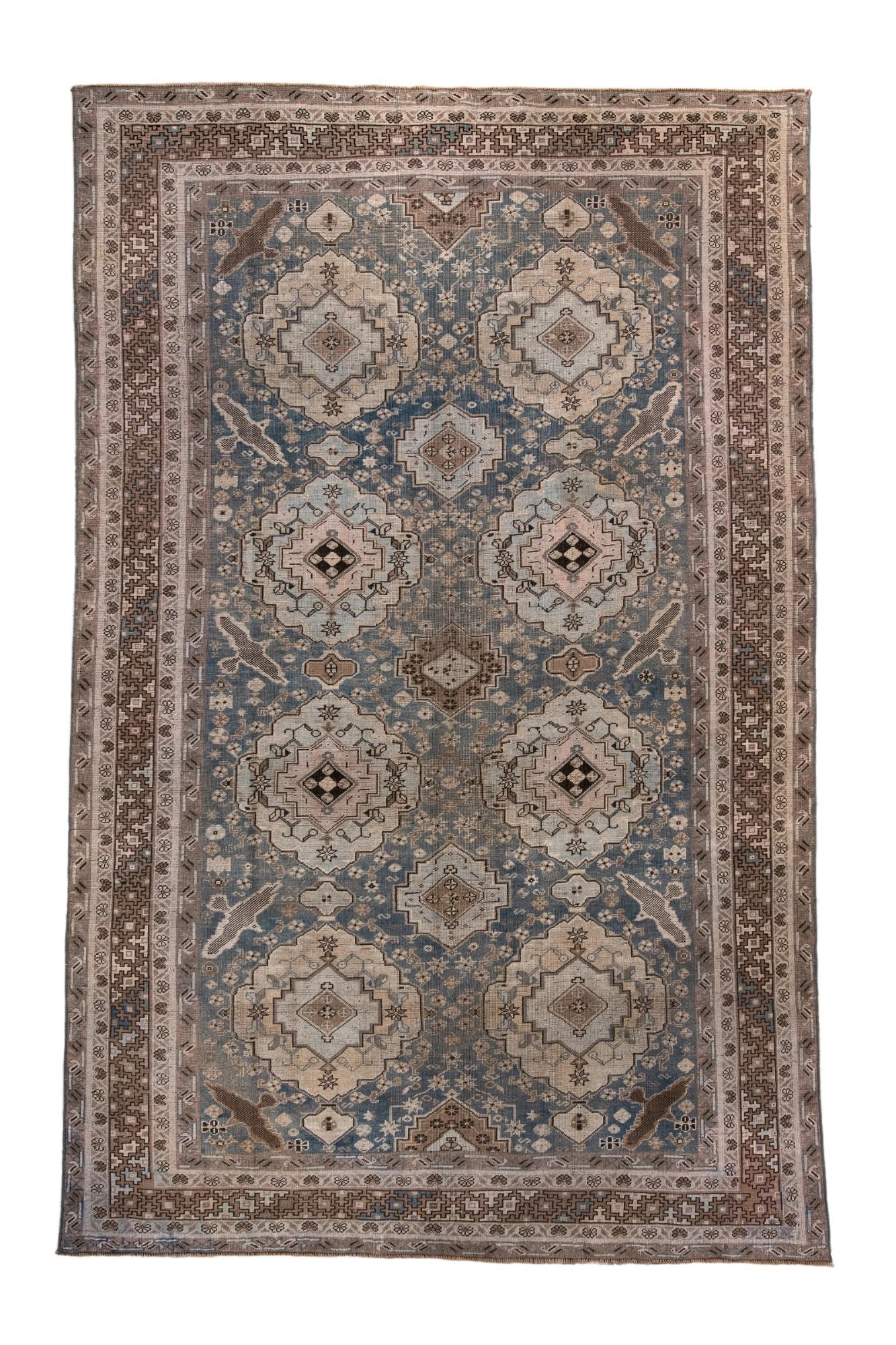 This antique Caucasian rug features a repeating series of geometric medallions on a soft blue field, framed by multiple tribal borders in taupe and ivory. Angular motifs, small rosettes, and classic Caucasian devices lend the piece strong decorative