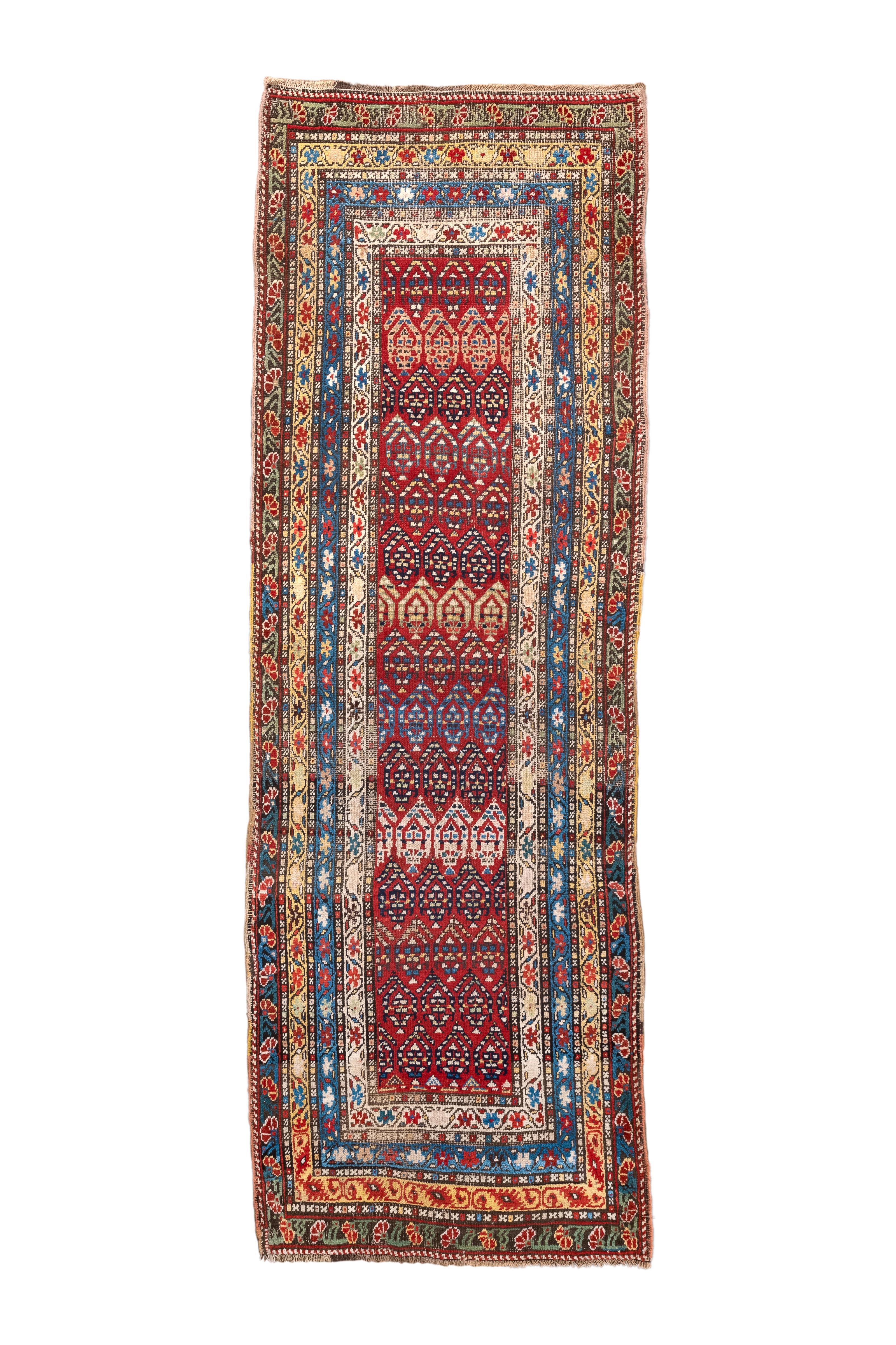 Ce kenare (coureur) Shulaver Kazak présente des botehs étroitement ajustés, décalés et inversant les rangs sur un champ rouge. Les botehs sont ornés de bandes colorées en bleu, crème, crème, marine et paille. Les bordures étroites présentent des