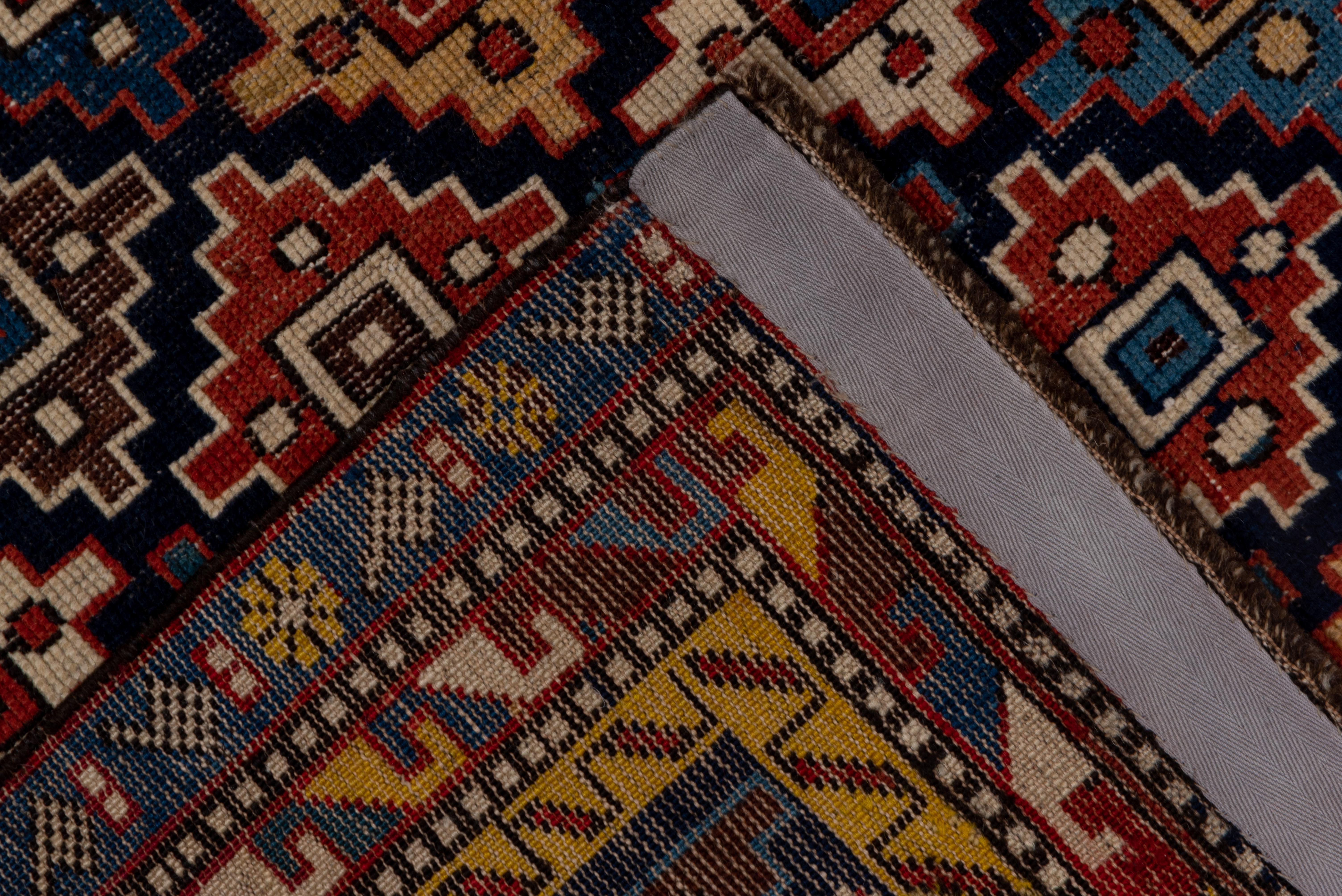 This boldly colored east Caucasian scatter rug shows a pattern of closely set, randomly toned stepped diamonds on a dark indigo ground. Red, cream, teal, blue shades and yellow are among the detail hues. Narrow borders centered on a polychrome