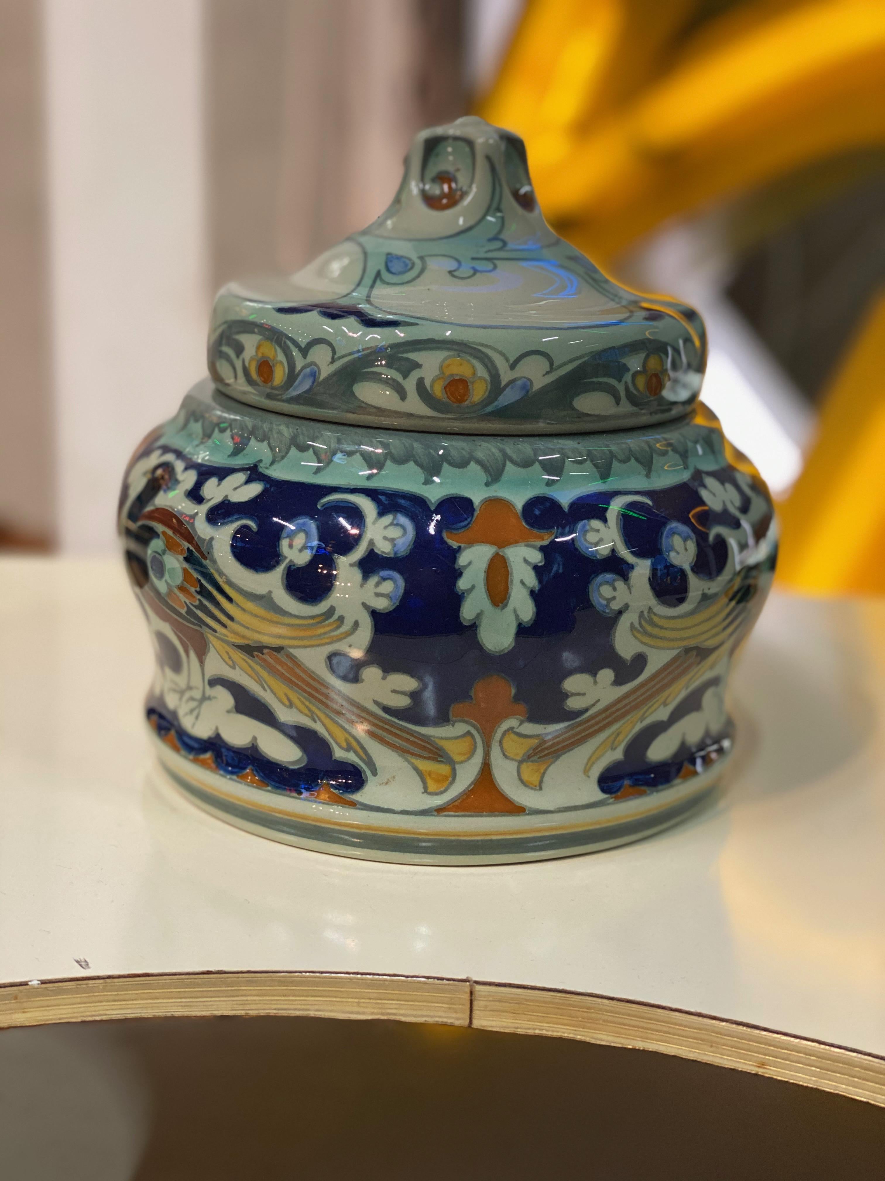 This beautiful and flawless little ceramic lidded box was designed by Samuel Schellink for N.V. Haagsche Plateelbakkerij Rozenburg. 
The lidded box in light grey was hand painted in several colors of blue, yellow, green and red. It shows a colorful