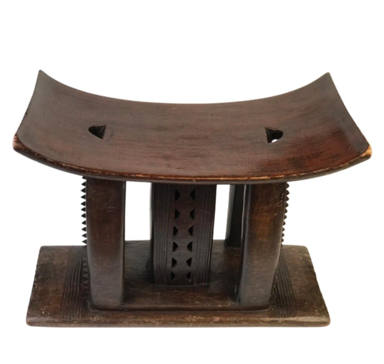 Ein antiker Ashanti Zeremonienhocker, aus einem einzigen Stück Holz geschnitzt. Der nach oben gewölbte Sitz steht auf fünf Stützen, von denen die mittlere mit kleinen Halbkreisen durchbrochen ist und  die vier äußeren Stützen sind geschnitzt und mit