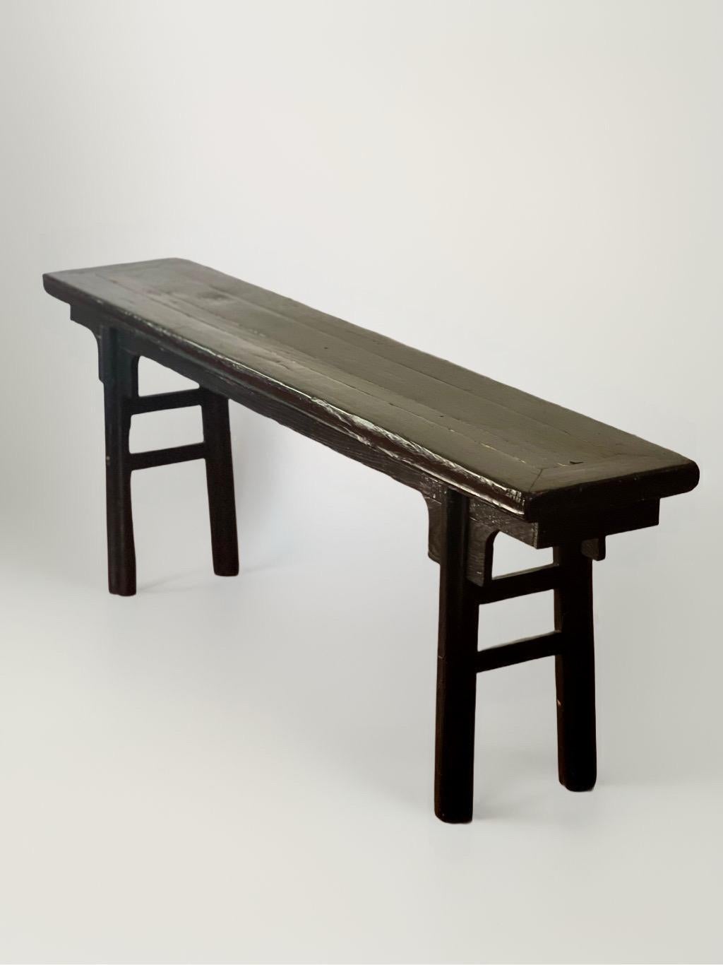 Antique primitive Chinese mortised black painted and lacquered bench from the early to mid 19th century. Simple in design with robust round legs, carved spandrels, clean lines and a weathered patina revealing undertones of original red paint through