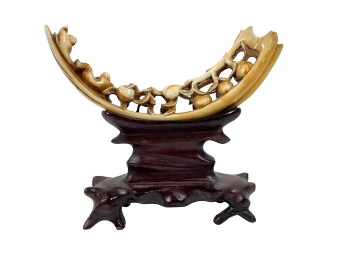 Chinese carved boar's tusk on carved hardwood stand. The tusk carved along the inner curve with conforming openwork peach branches with leaves and peaches and extending to two men reclining at the tip. Set on a carved hardwood stand conforming to