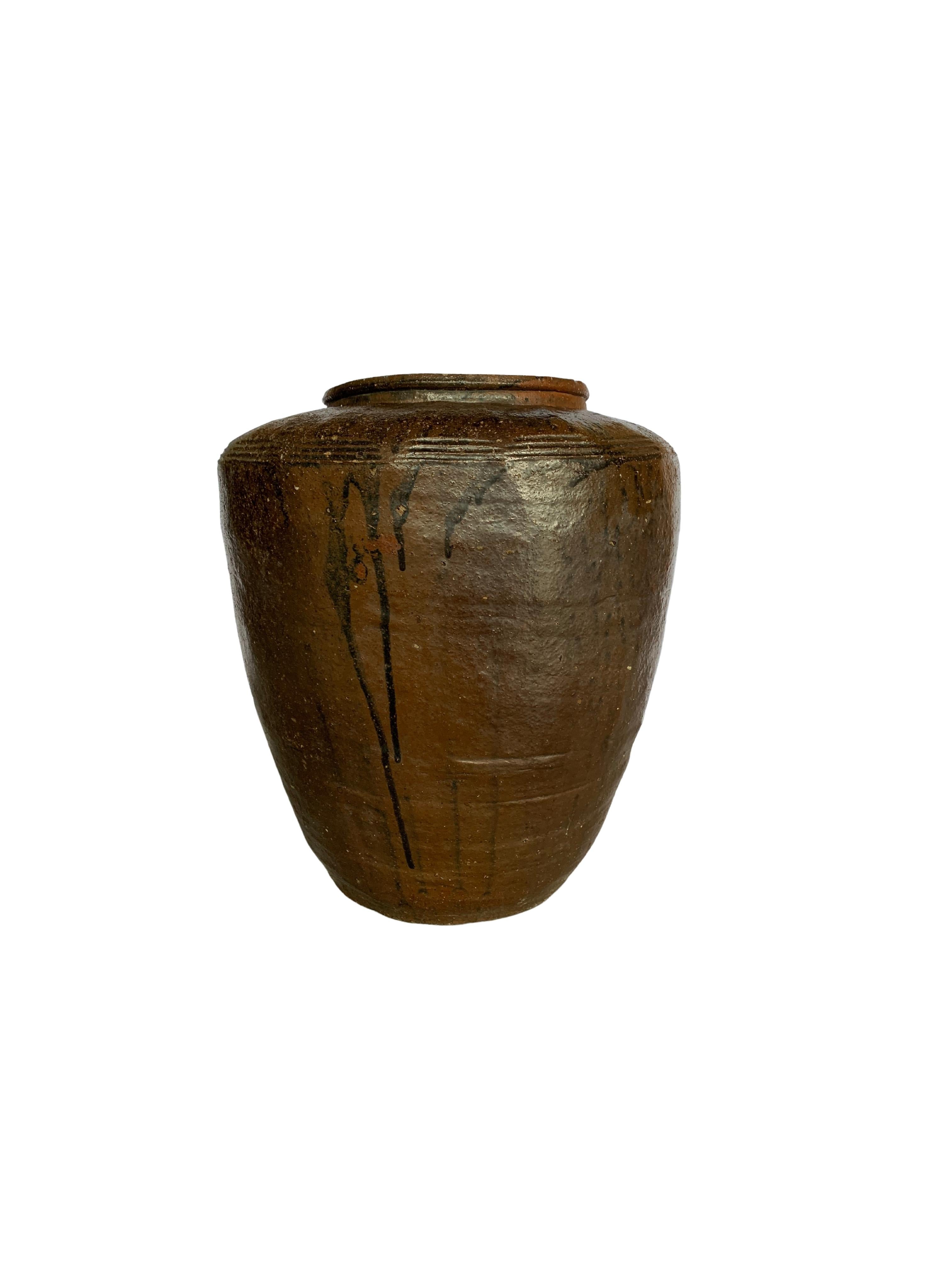 This Antique Jar dating to the early 1900s once served the purpose of crafting salty eggs. The jar features a wonderful brown glaze with some areas of dripping black. There is a ridged pattern finish near to the Jar's rim. The fading and markings to