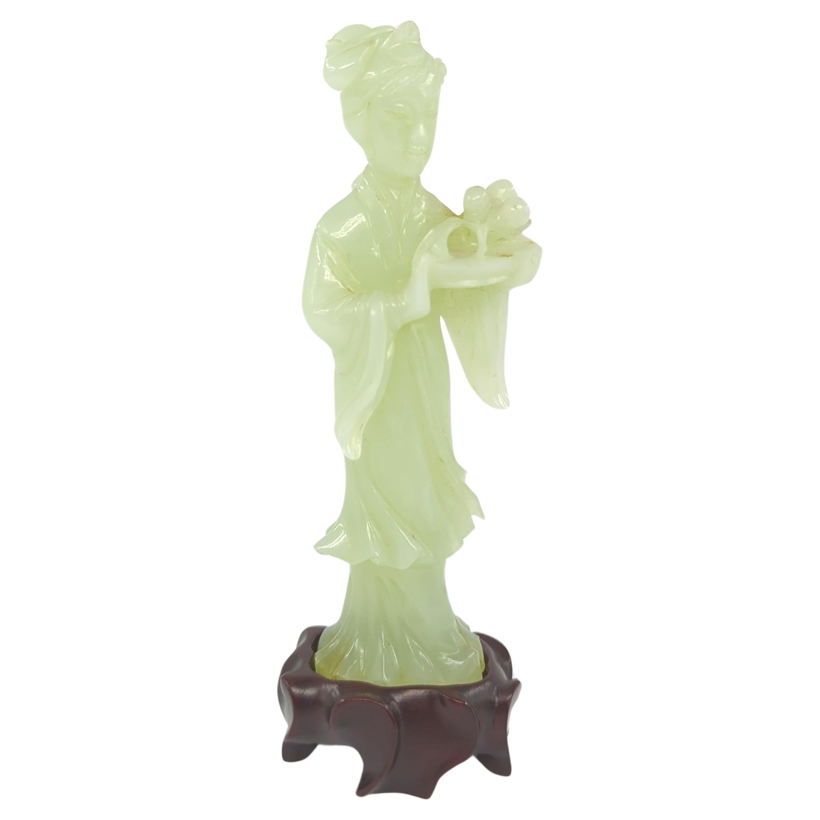 This masterfully carved celadon jade figure represents the epitome of Chinese artistry and craftsmanship. The sculpture features a beauty dressed in flowing traditional attire, capturing the essence of grace and elegance. She is depicted holding a