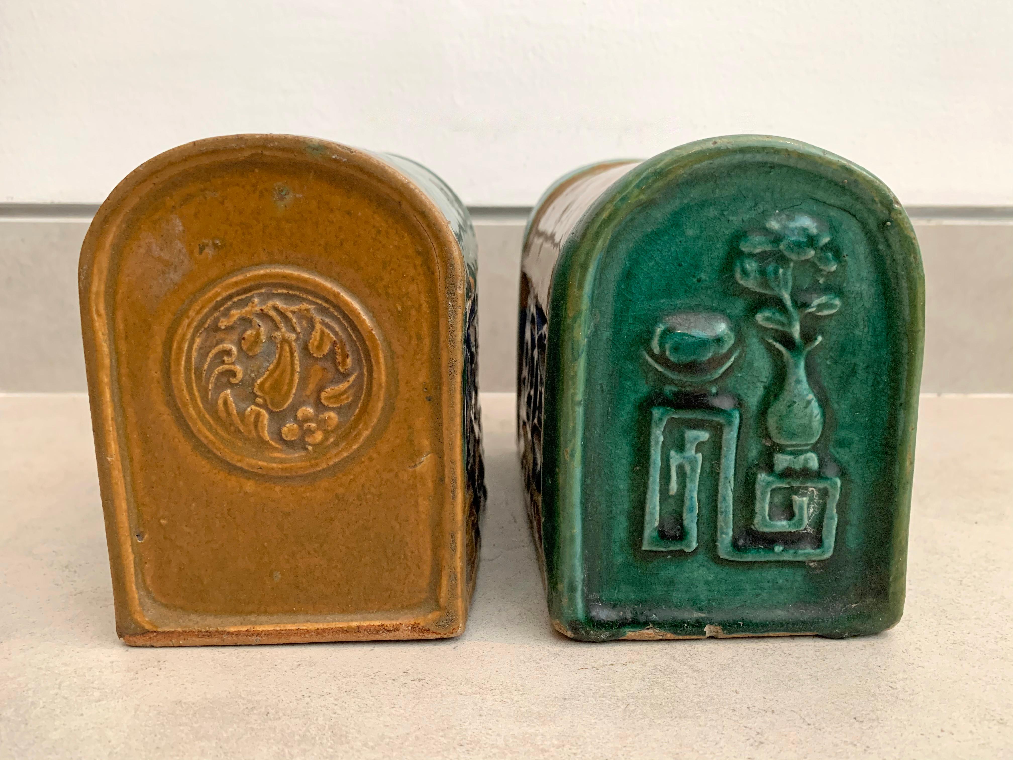 This pair of Chinese ceramic headrests feature a green, brown and blue glaze as well as floral motifs. They also feature pierced sides with symbols of Chinese coins which are a symbol of wealth. 

They differ slightly in size: height 13cm x width