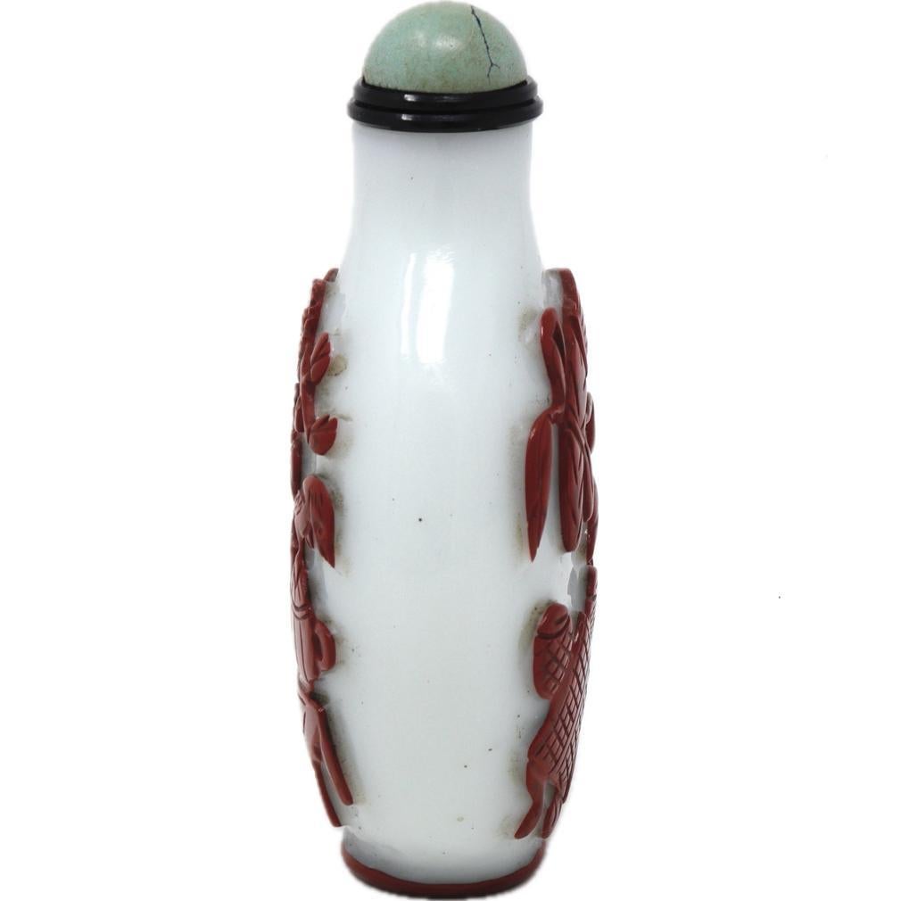 A Cinnabar-red Overlay White Glass Snuff Bottle, 19th century. The flat oval white glass bottle has a thinn overlay of opaque dense red glass resembling cinnabar lacquer and deeply carved away in a cinnabar style to reveal a scholar’s studio with a