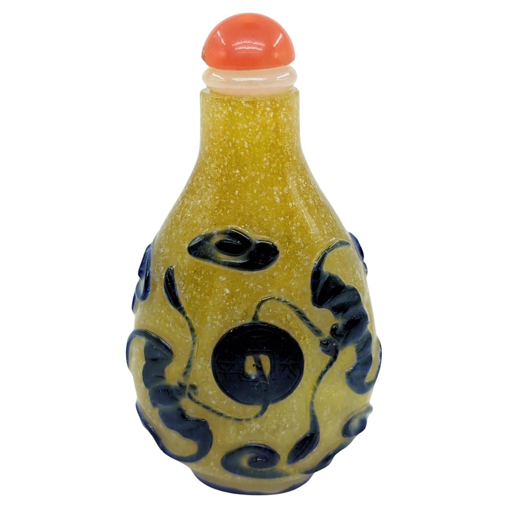 This fine Qing period antique snuff bottle is a beautiful example of color glass overlay craftsmanship. It features a vibrant yellow snow storm ground, over which dark blue glass has been overlaid to create a striking design. The central motif on