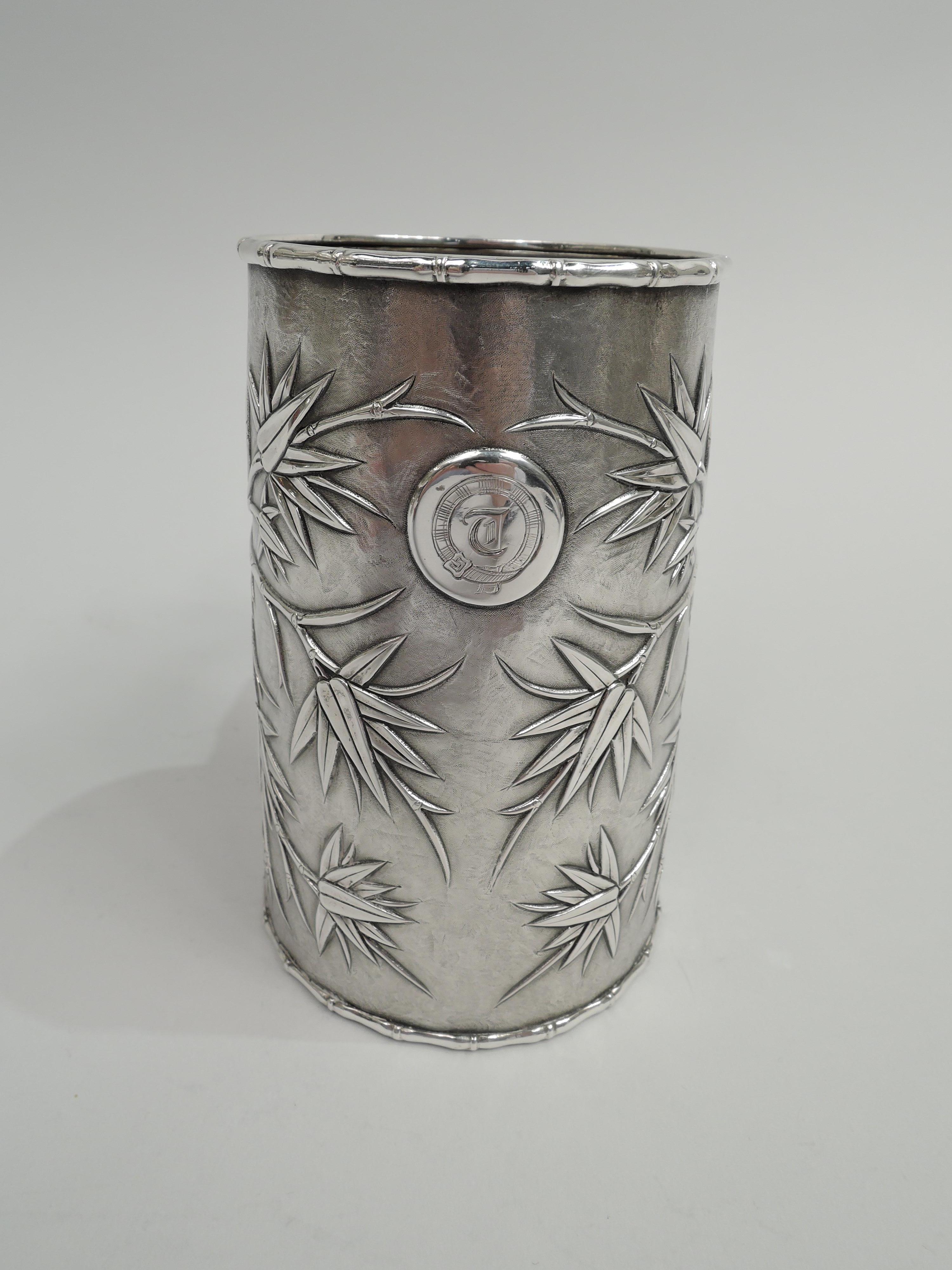 Chinese export silver mug, ca 1850. Straight and upward tapering sides applied with leafing bamboo on stippled ground. Branch-form handle and rims. Applied rondel engraved with single-letter monogram in buckled belt. On underside is engraved “H. J.