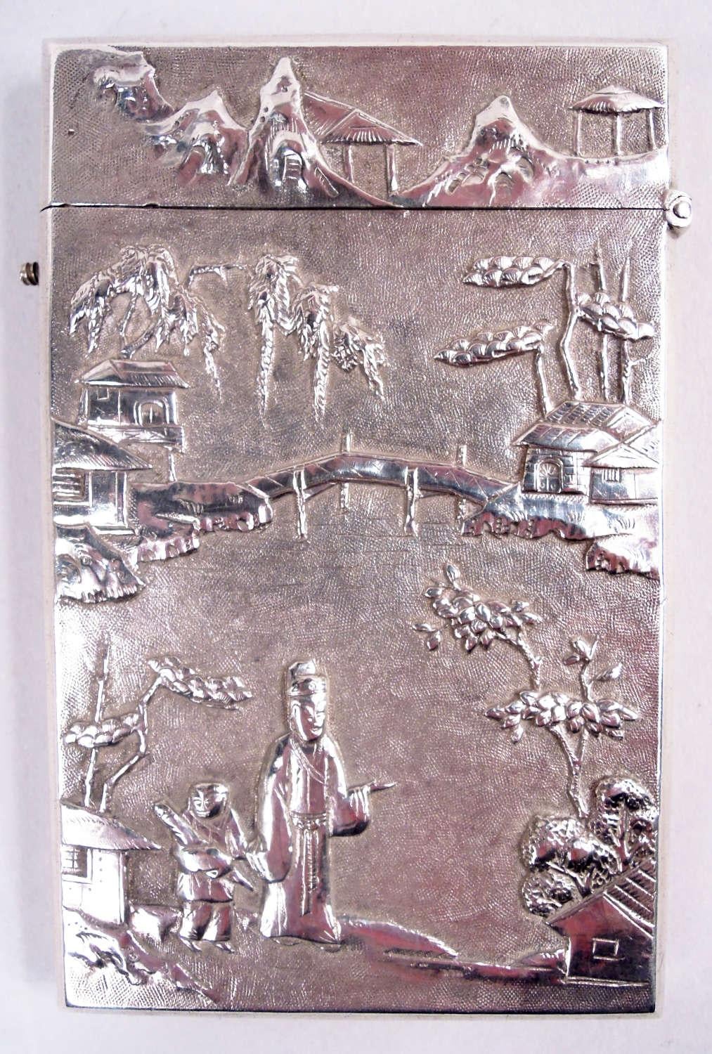 Chinese export silver card case, ca 1910. Rectangular with hinged push-button cover. Chased ornament on stippled ground. On recto, a blossoming prunus branch and birds with armorial cartouche (vacant). On verso, a mountainous landscape with bridges