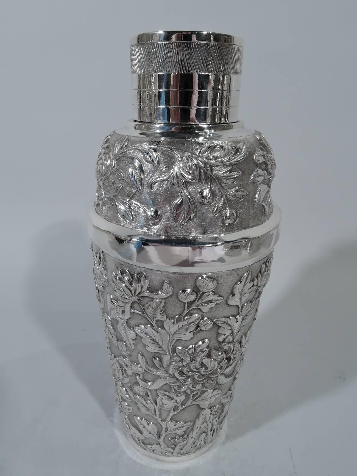 Chinese export silver cocktail shaker, circa 1900. Tapering cup. Cover has curved shoulders and cylindrical neck with cap and built-in strainer. Repousse chrysanthemums on stippled ground. Cover sides have engraved bands and dense slash lines.