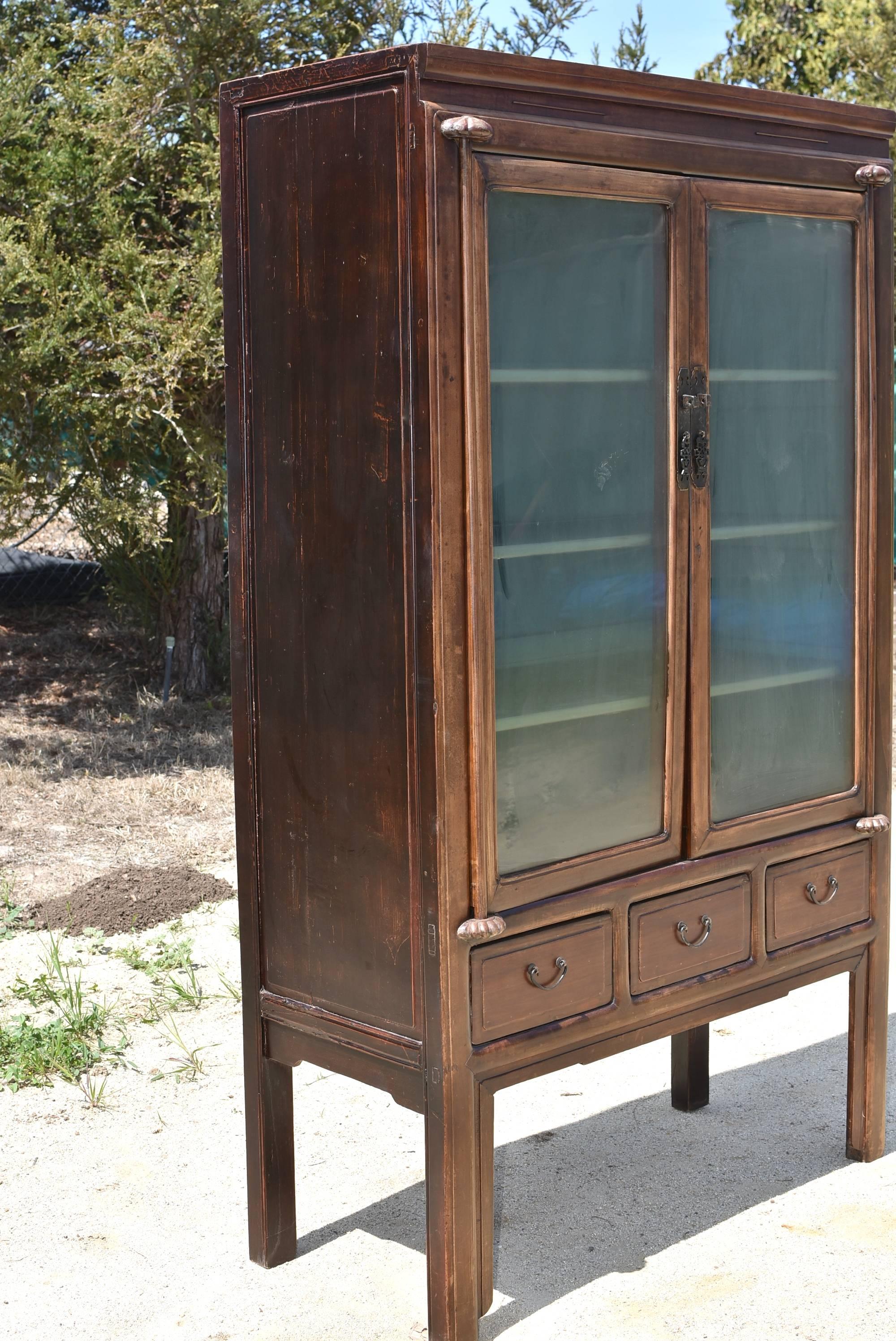 A Ming style solid wood cabinet with glass doors and removable shelves. Three full length drawers provide additional storage. Carved thin lines on the top reflect Ming influence. Beautiful hardware is of a flower basket pattern. Half lotus knobs
