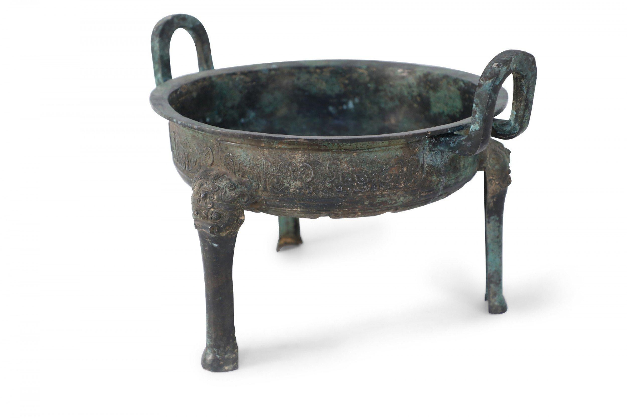 Antique Chinese Han Dynasty-style copy of a patinated bronze pot carved with raised geometric patterned bands wrapping its form, sitting atop three legs, and fitted with two upright handles.
  