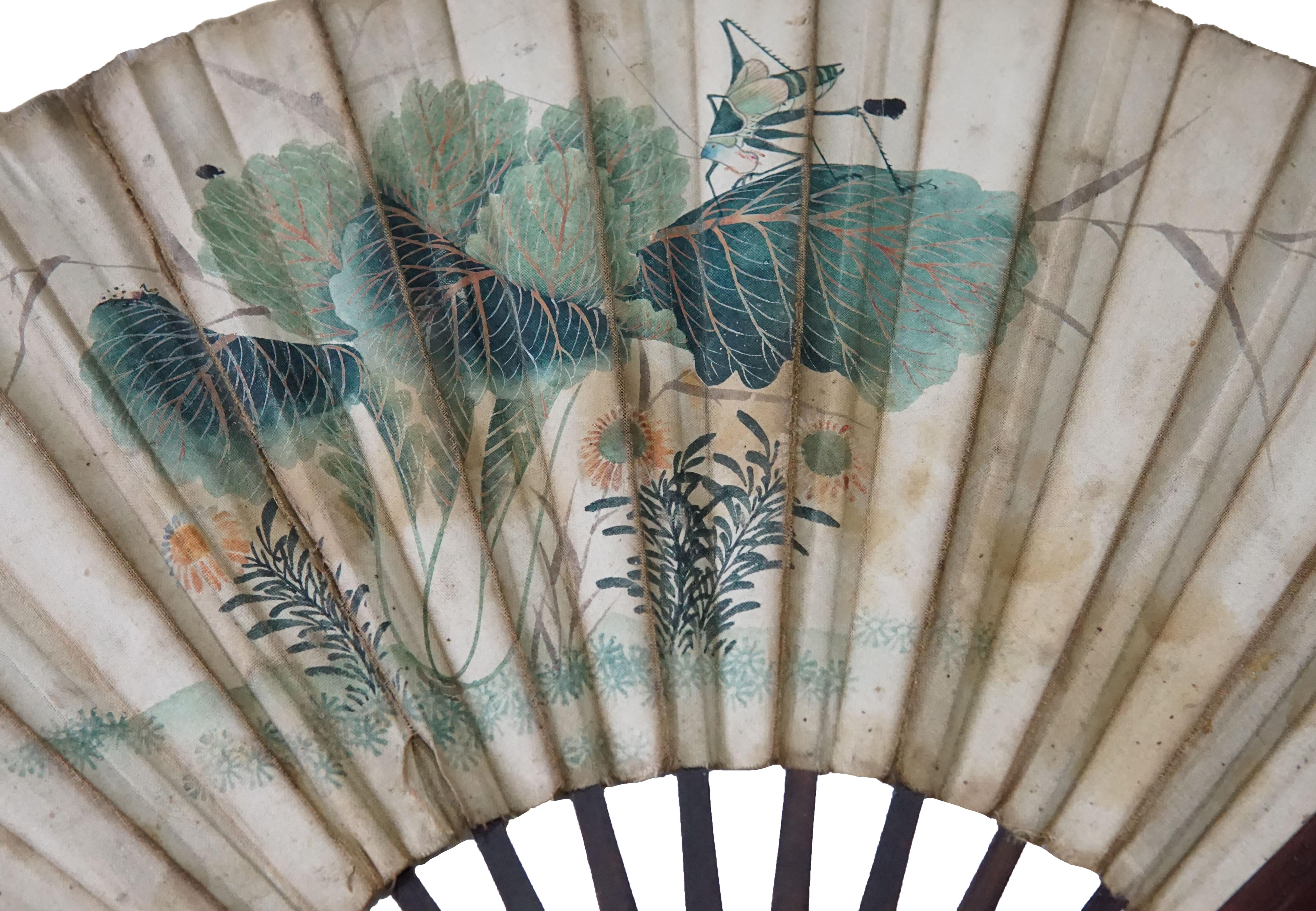 A Chinese hand-painted fan with hand-painted calligraphy (with two different drawing on either side). The fan's frame is crafted from bamboo and also includes wood engravings. The calligraphy features flowers, leaves and a grass hopper on one side &