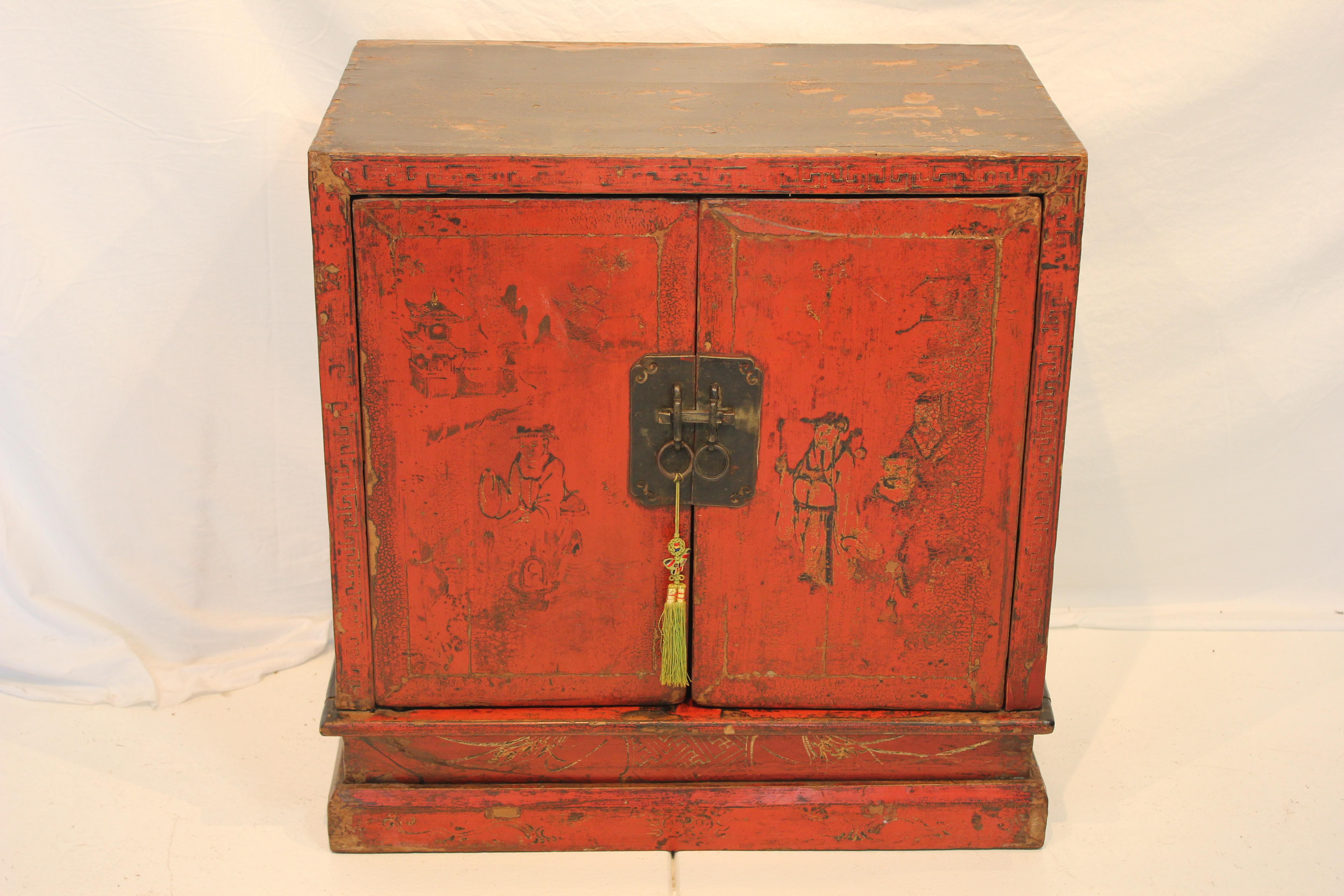 Mid 19th Century Chinese Hand Painted Red Lacquer Chinoiserie Two Door Cabinet, having a Chinoiserie gilt painted scene on the rectangular top and two hinged door panels. Pair of brass lifting handles on side panels, brass lock plate and pin