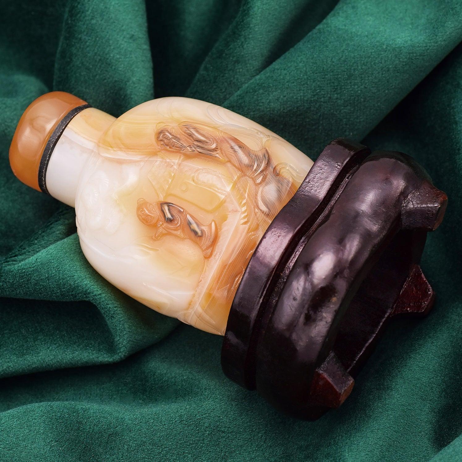 Antique Chinese snuff bottle finely carved from light-toned agate, featuring an intricate landscape scene with figures and a house in relief. It is topped with a warm orange stopper and displayed on a wooden base. A graceful example of traditional