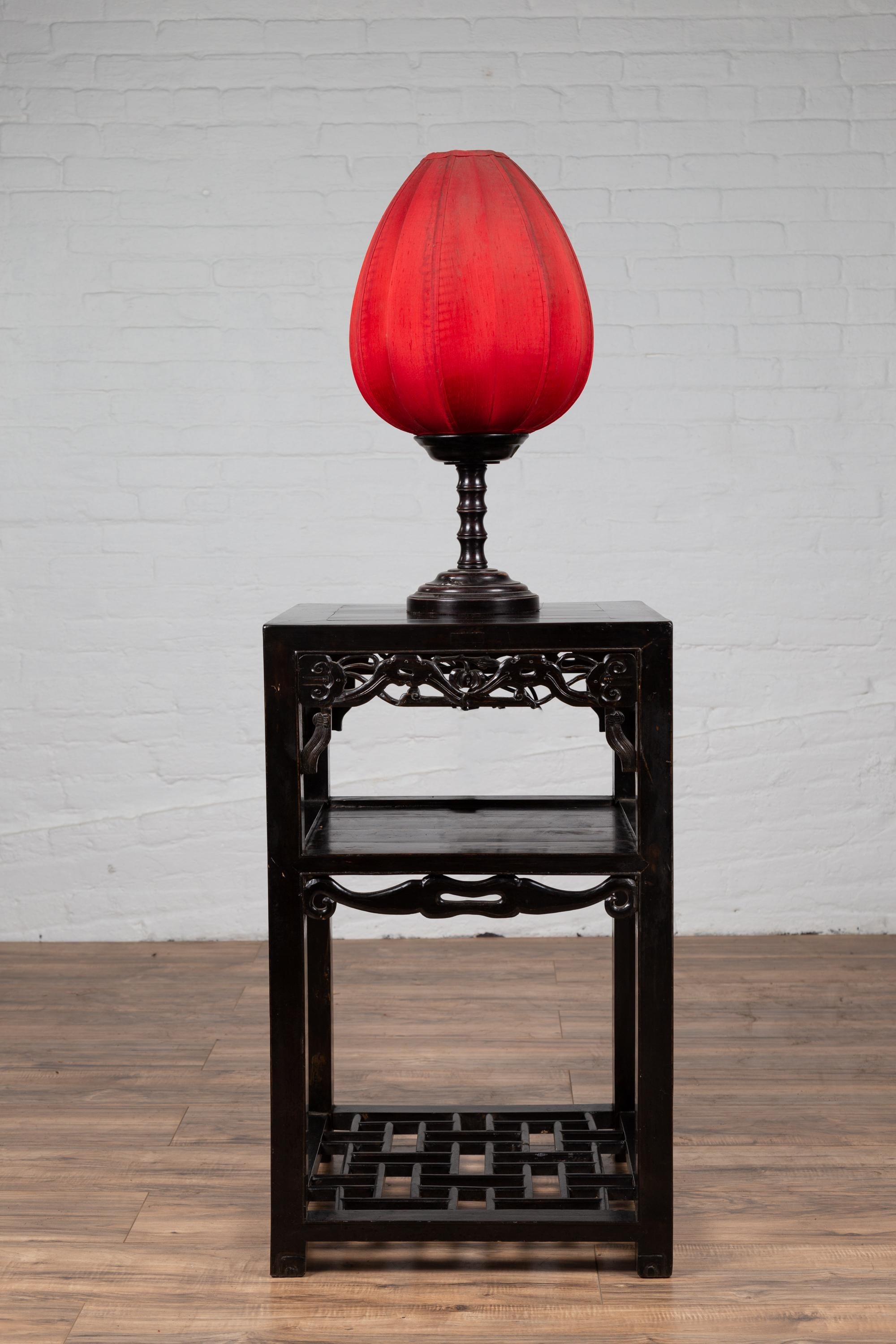 An antique Chinese lamp table from the early 20th century, with dark patina, carved apron and shelves. Born in China during the early years of the 20th century, this exquisite side table features a square top with central board, sitting above a