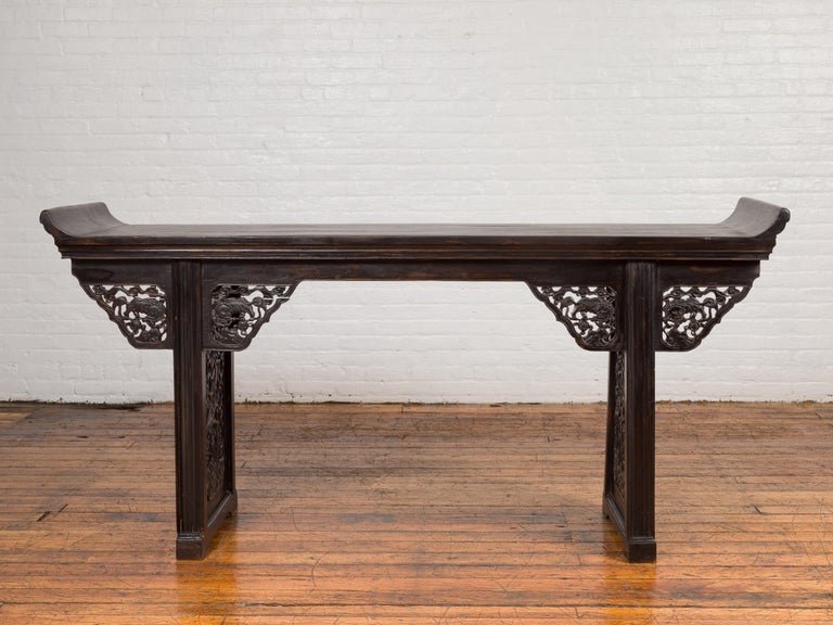 An antique Chinese Ming dynasty style altar console table from the 19th century, with black patina, everted flanges, carved spandrels and sides. Born in China during the 19th century, this altar console table features a rectangular planked top
