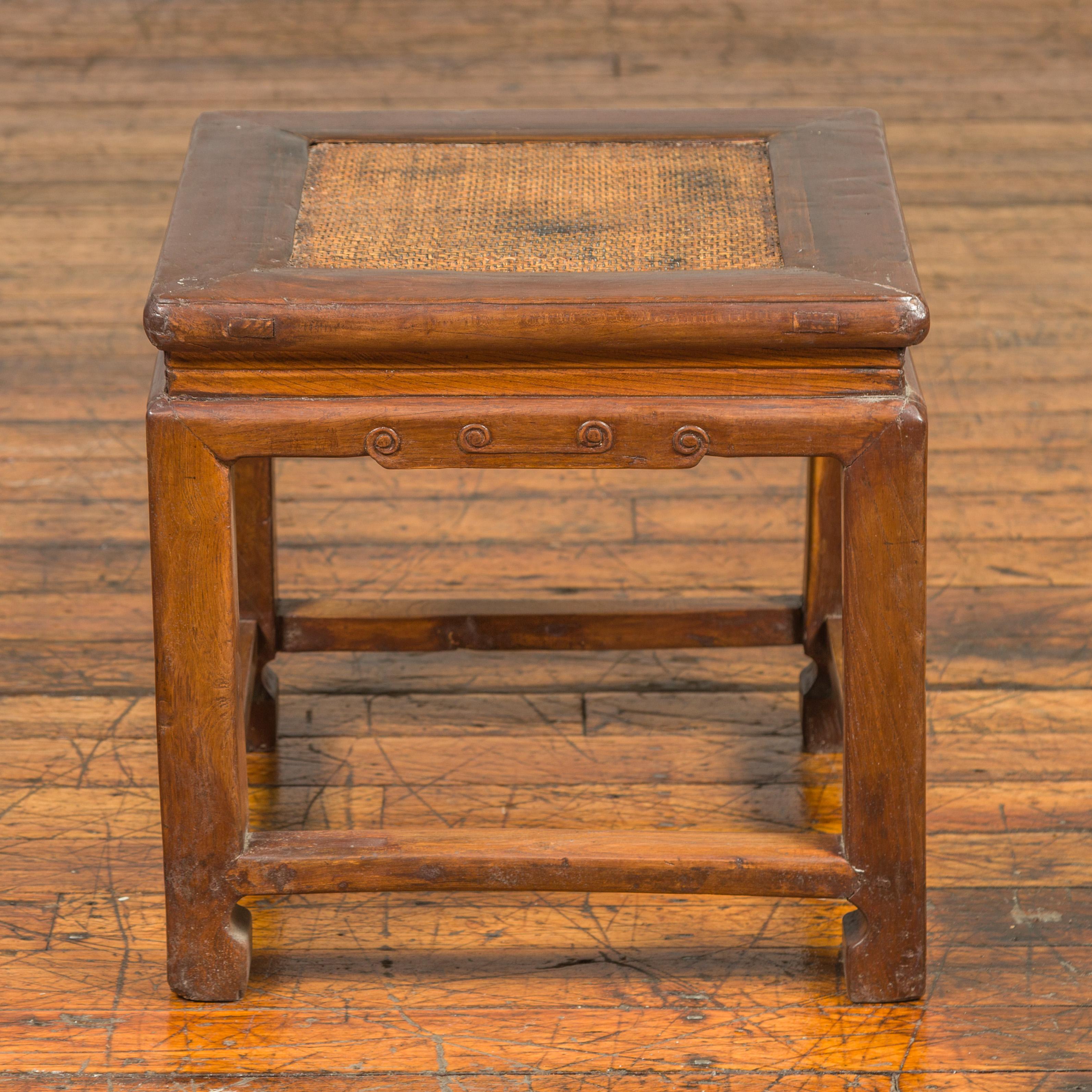 An antique Chinese Ming Dynasty style wooden waisted stool from the early 20th century, with rattan inset, horse hoof legs and cloud motifs. Born in China during the early years of the 20th century, this stool features a woven rattan square top