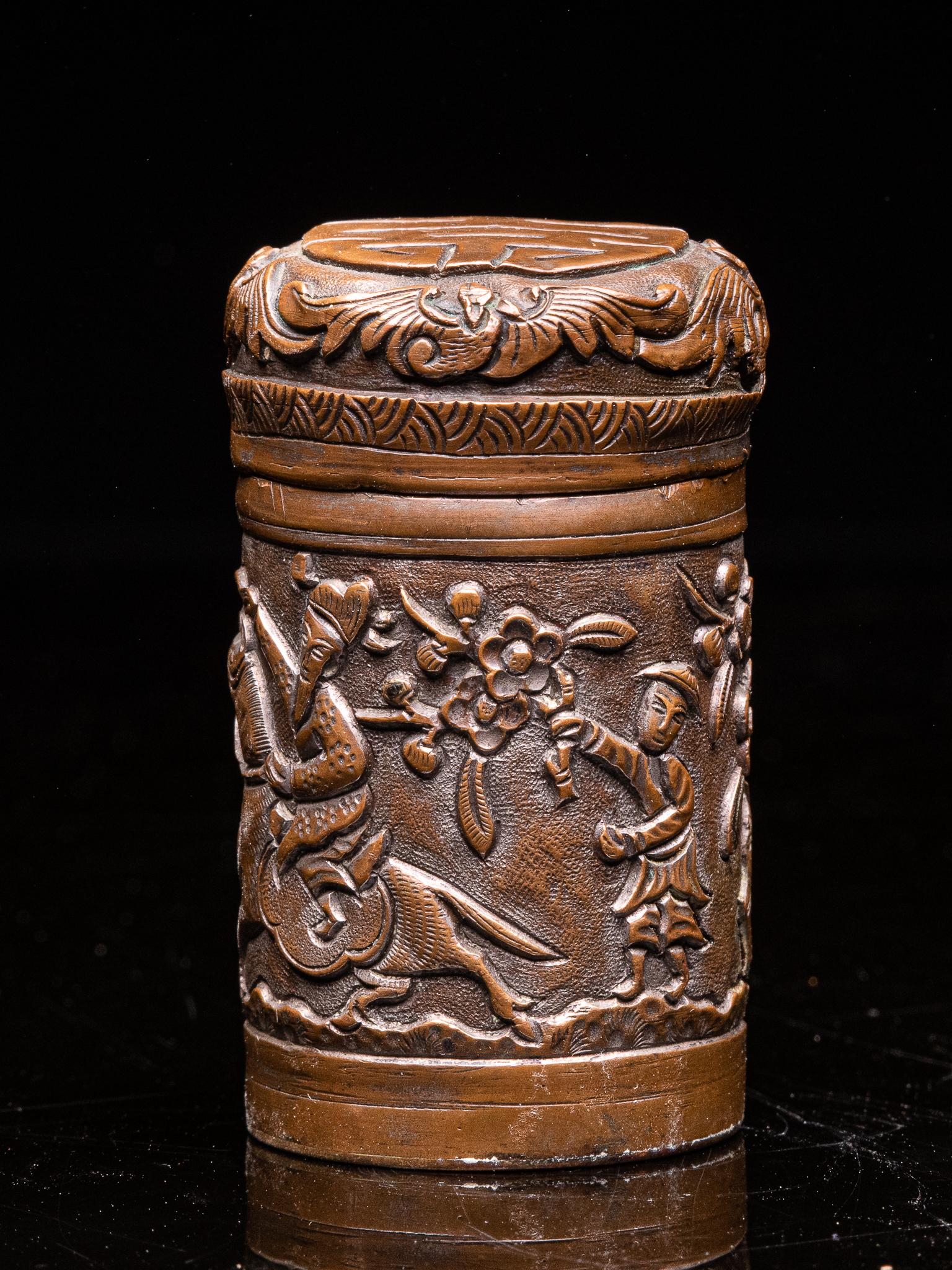 The cylindrical opium container is executed in hammered brass and decorated with an expressive repose design of a rider in a landscape with blossom trees and birds. The lid is decorated with a shou symbol, which refers to a long life.