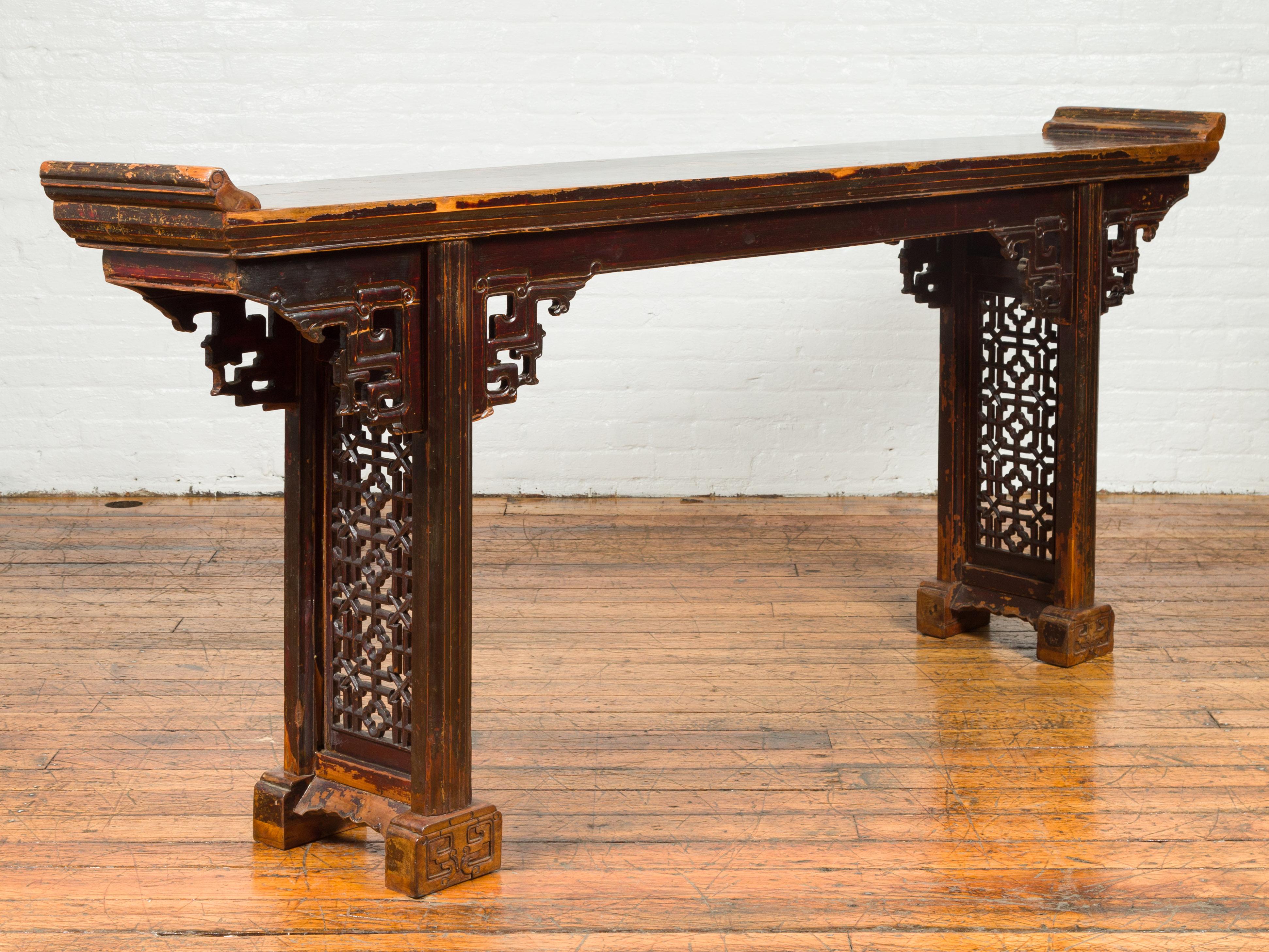 A Chinese Qing dynasty period altar console table from the 19th century, with dark distressed patina, everted flanges, carved spandrels and sides. Born in China during the Qing dynasty, this altar console table features a rectangular planked top
