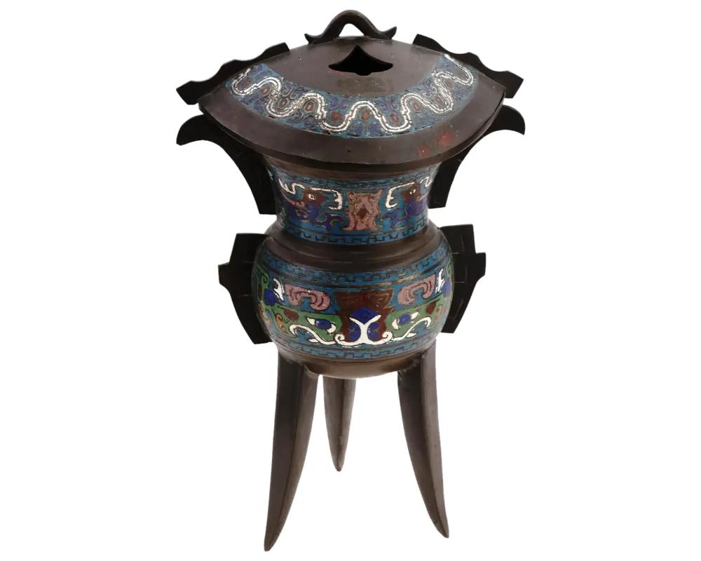 An antique Chinese copper tripod Jia vessel with a single handle. Qing Dynasty, 1636 to 1912. Three conical legs hold lower bowl section. The upper tapered section is closed with a hinged lid. The body of the vessel is garnished with cloisonne