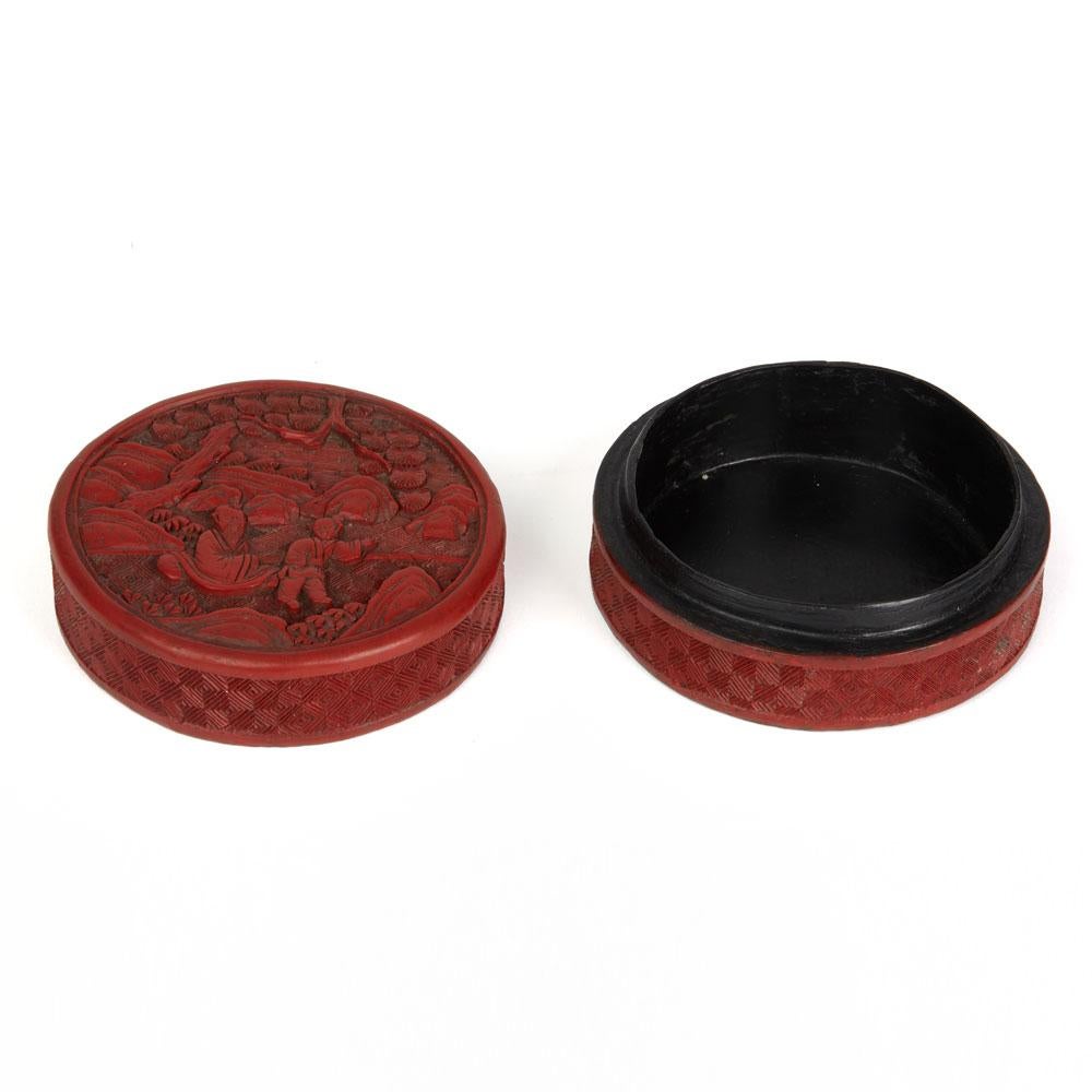 A fine antique Chinese red cinnabar lacquer lidded rounded box carved with a scene with a young boy and a seated elderly bearded sage within a rocky landscape with trees. The box has a fine diamond pattern design around the sides with a black