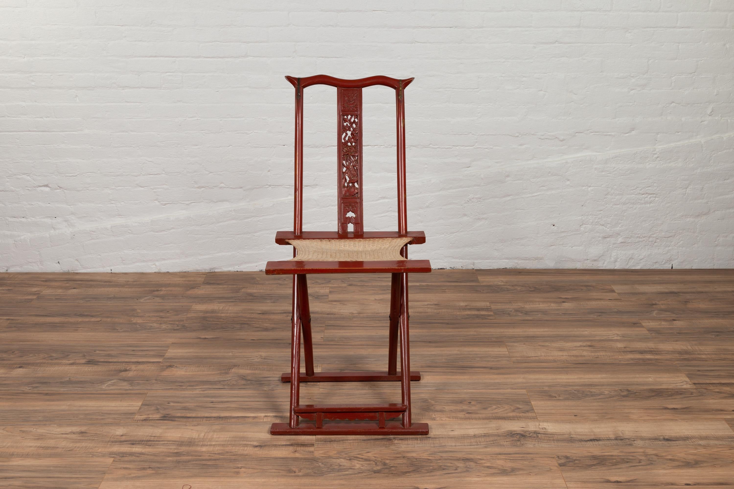 A Chinese late Qing Dynasty period red lacquered folding traveller’s chair from the early 20th century, with carved motifs, footrest and woven fabric. This early 20th-century antique Chinese traveler's chair is a striking example of functionality
